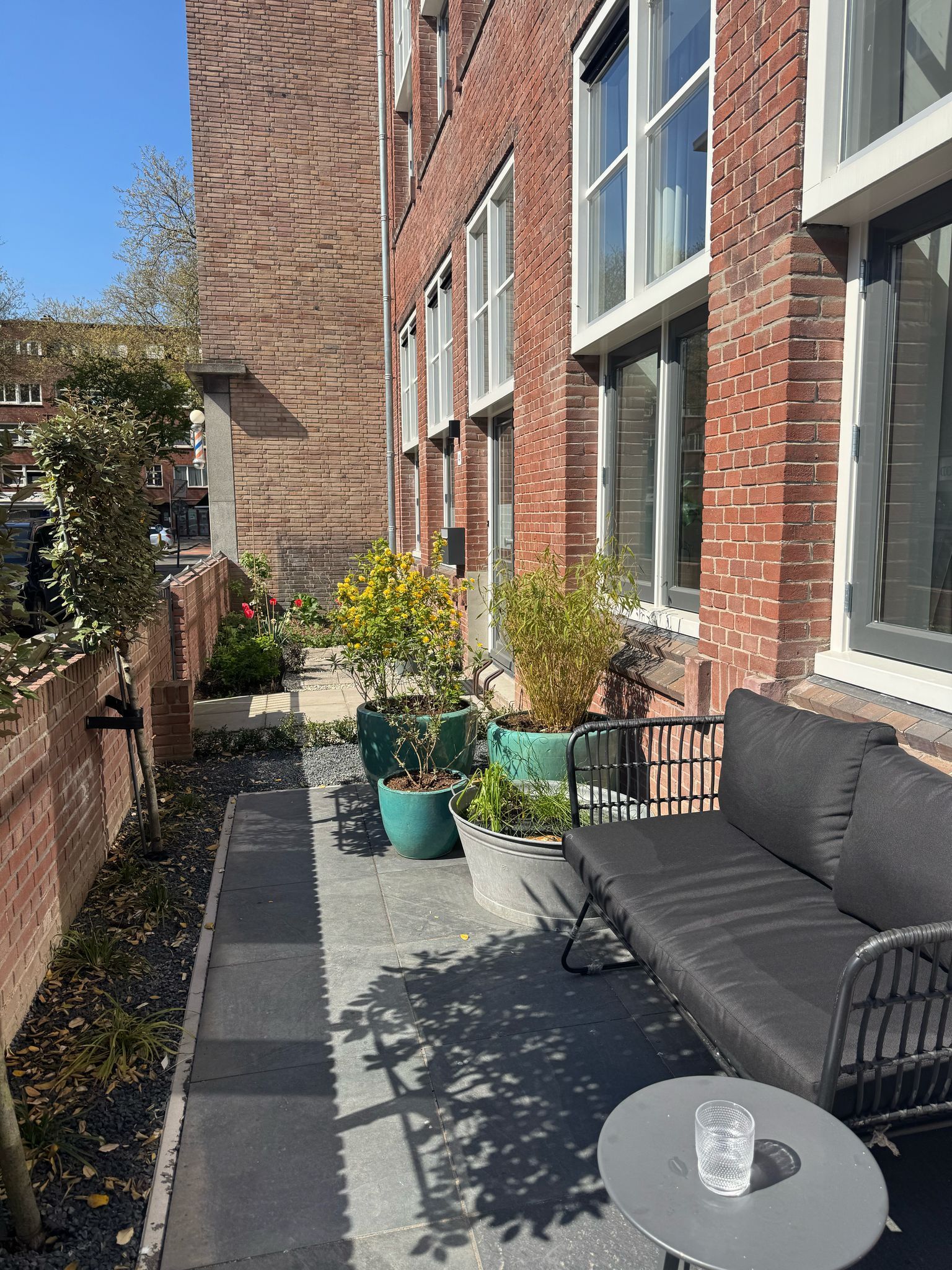Smal terras met zitgelegenheid, potplanten en een rood bakstenen gebouw. ​​Zonnige dag.