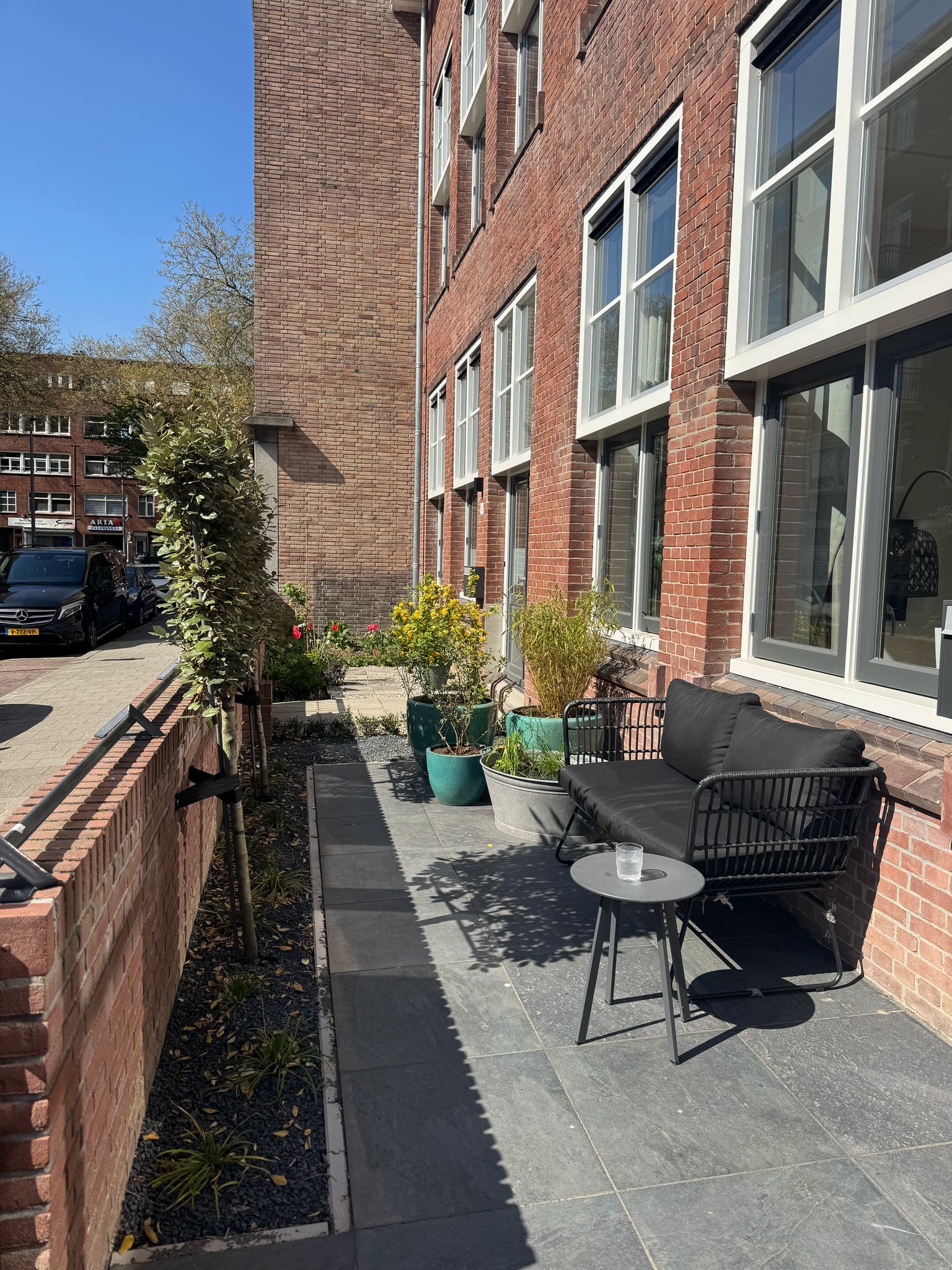 De buitenkant van een bakstenen gebouw met zitgelegenheid buiten en potplanten op een grijs terras. Zonnige dag.
