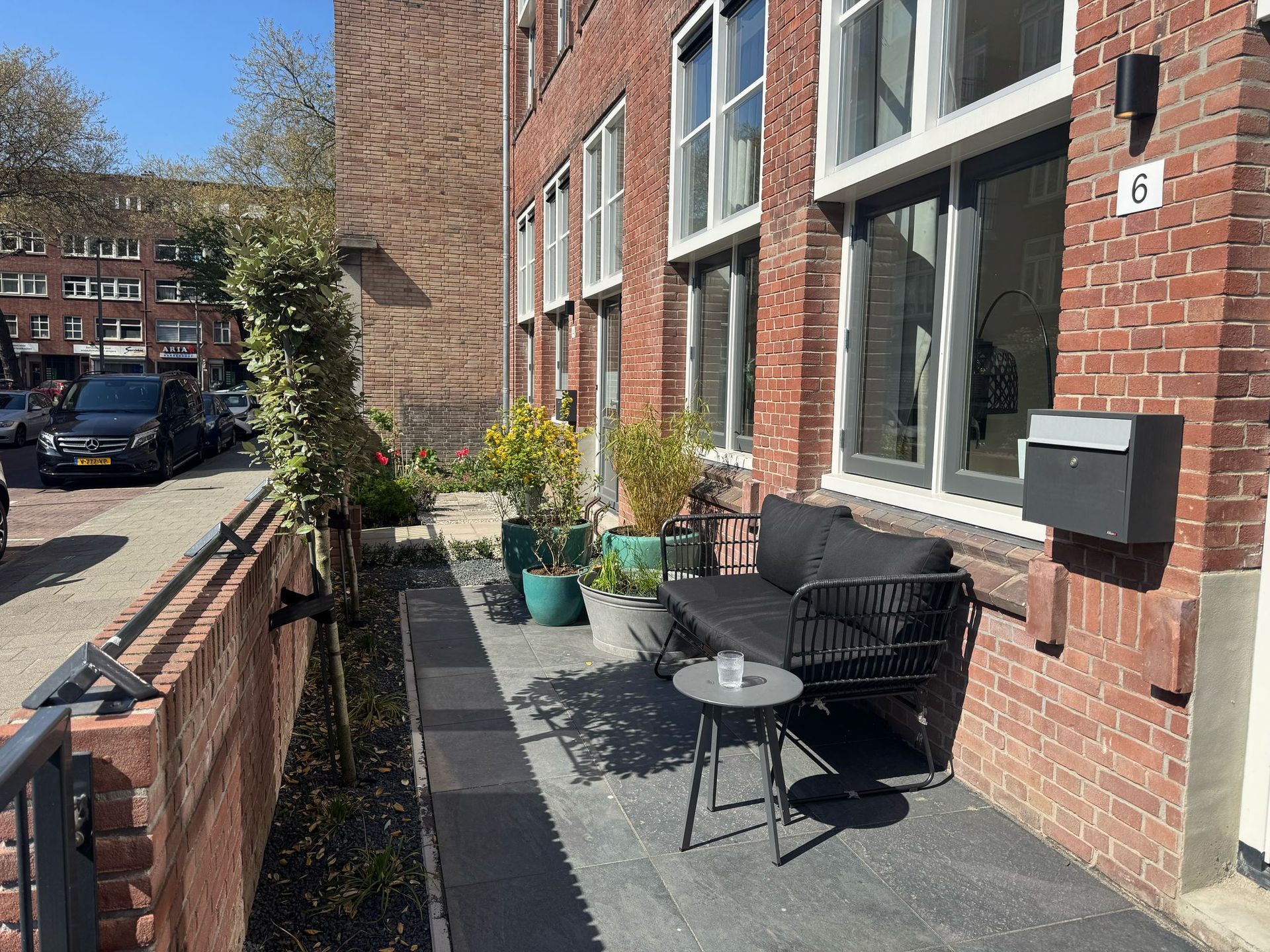De buitenkant van een bakstenen gebouw is voorzien van een terras met zitgelegenheid, potplanten en een zwarte brievenbus.