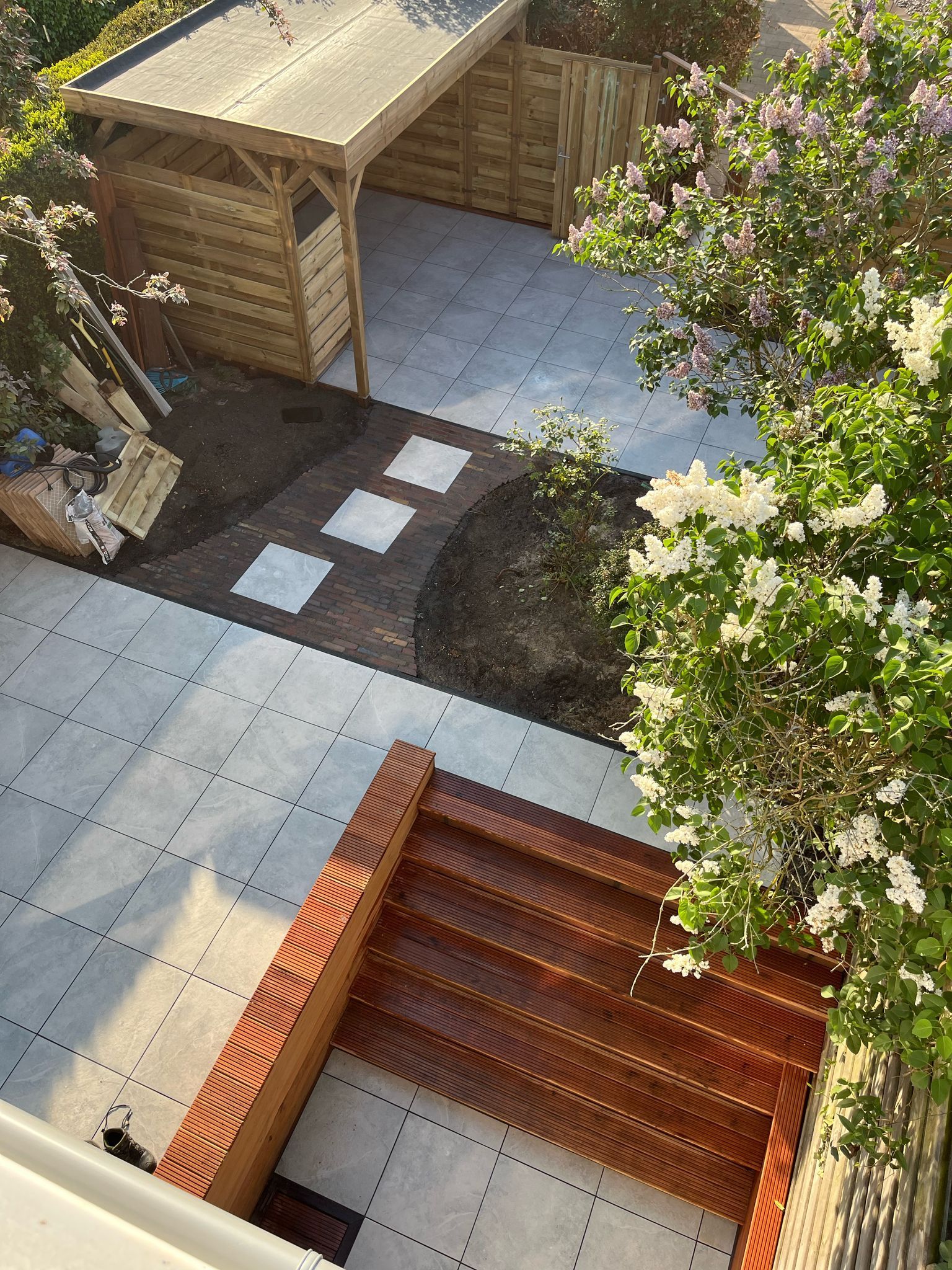Een patio met een houten carport, bestrate paden en bloeiende seringenstruiken.