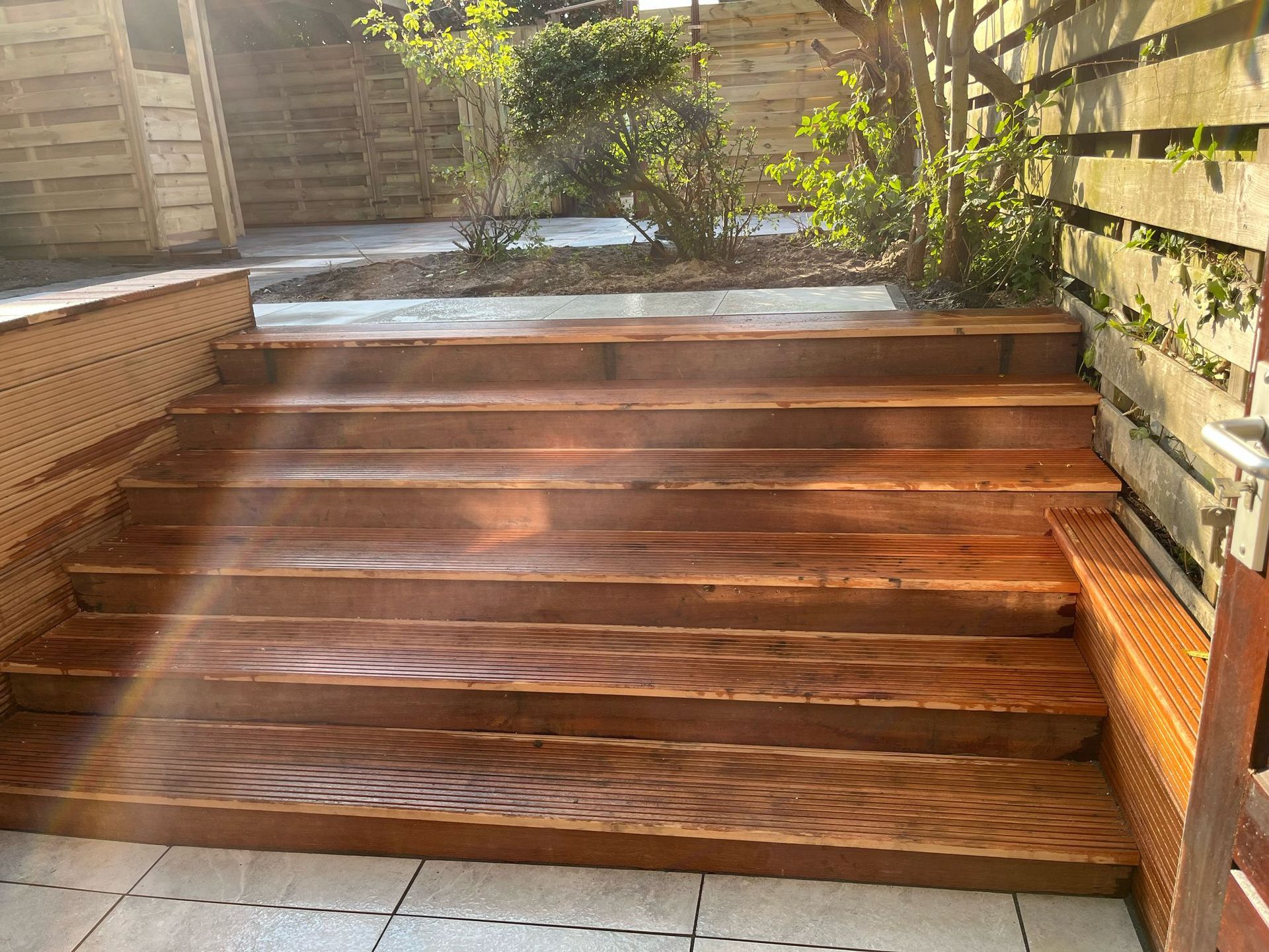 Houten buitentrappen leiden naar een tuin; zonlicht stroomt erdoorheen.