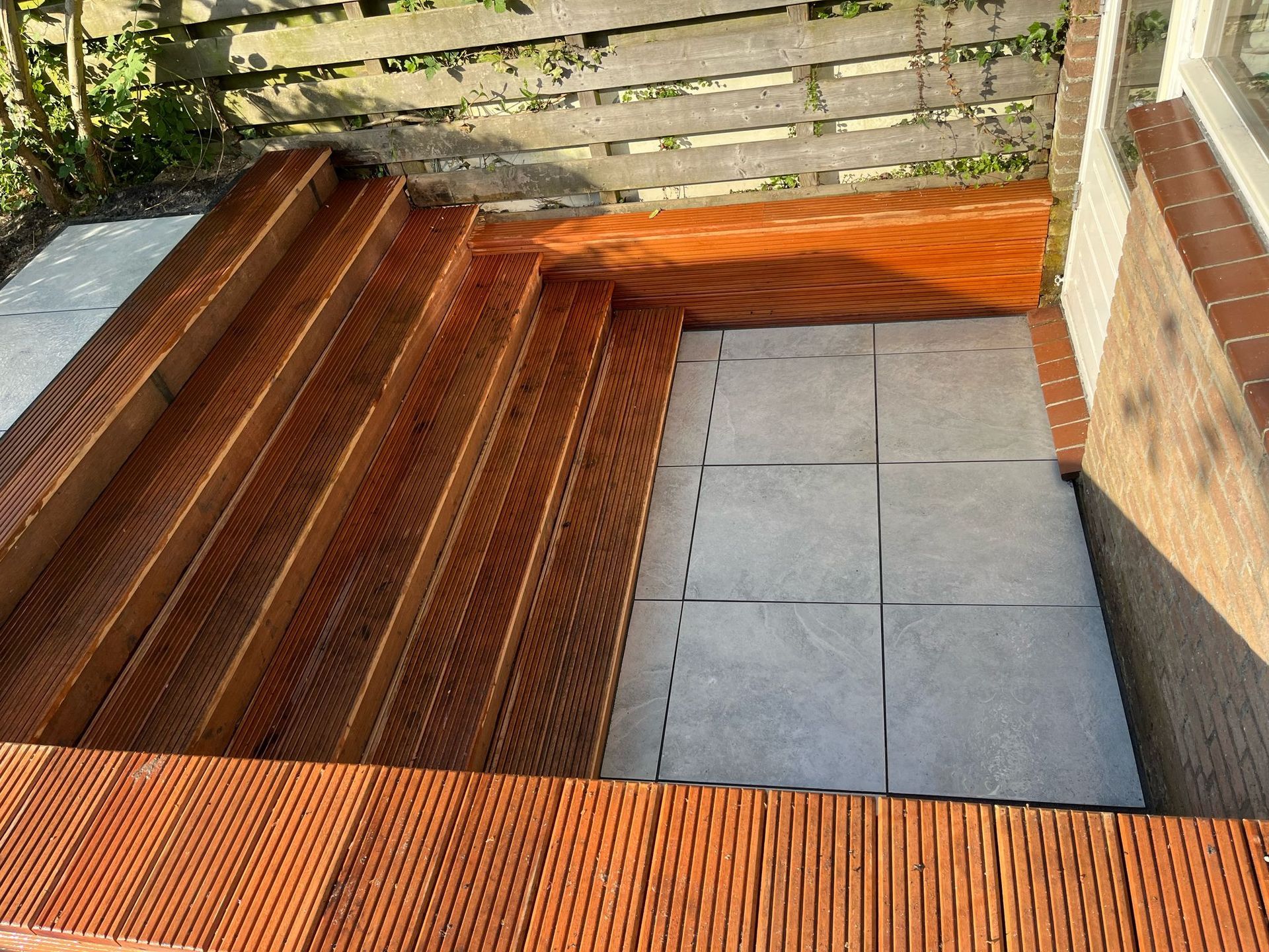 Een houten trap leidt naar een terras met grijze tegels, naast een bakstenen muur en een hek.