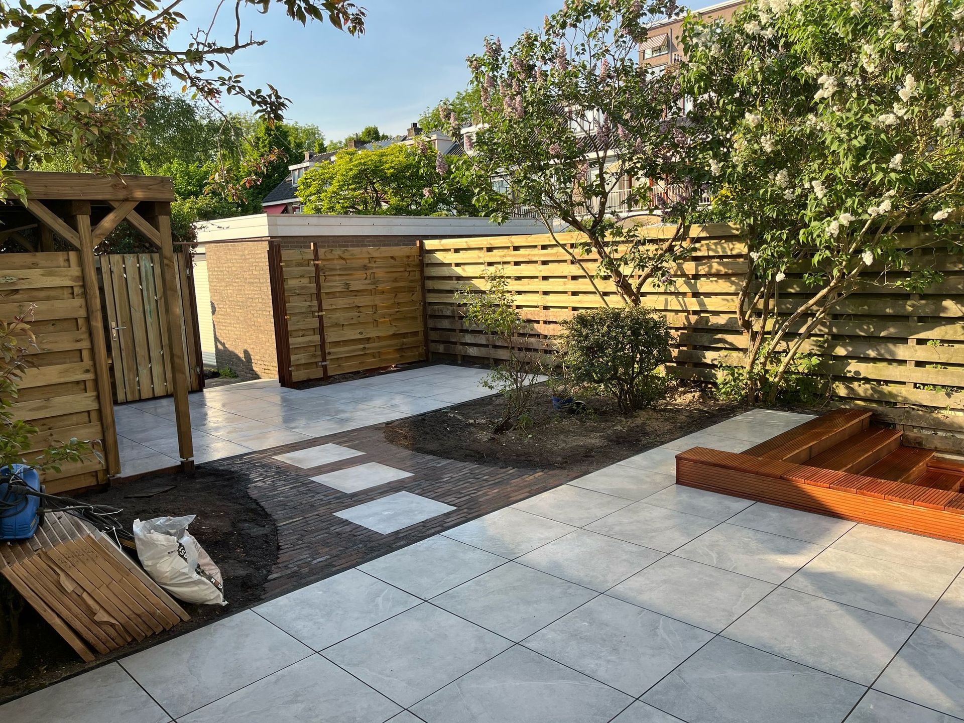 Een terras in de achtertuin met tegelvloer, houten schutting, een smal pad en een zandbak.