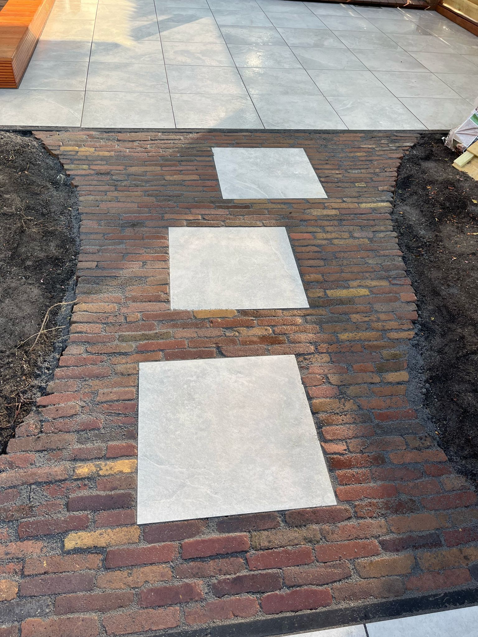 Een bakstenen pad met lichtgrijze vierkante stapstenen leidt naar een betegeld terras.