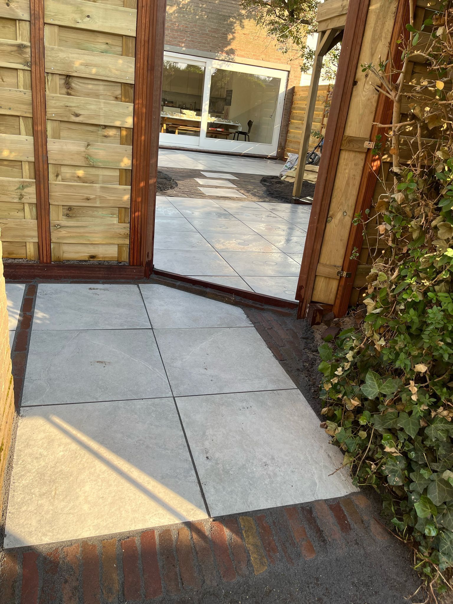 Een pad met straatstenen leidt naar een gebouw met een wit omlijste deuropening, omgeven door een houten hek.