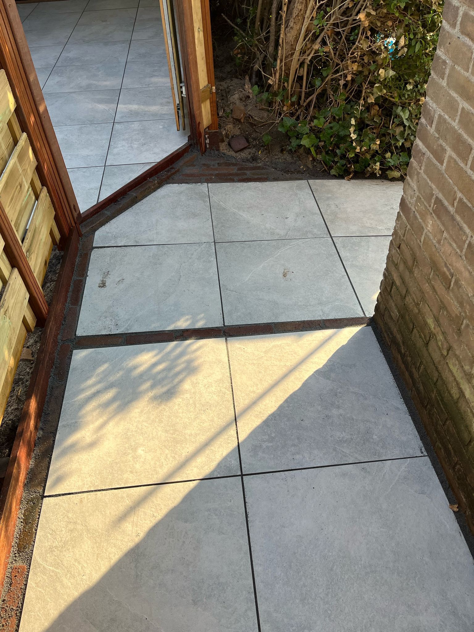 Betonnen terras met donkere lijnen, bruine schutting en bakstenen muur.
