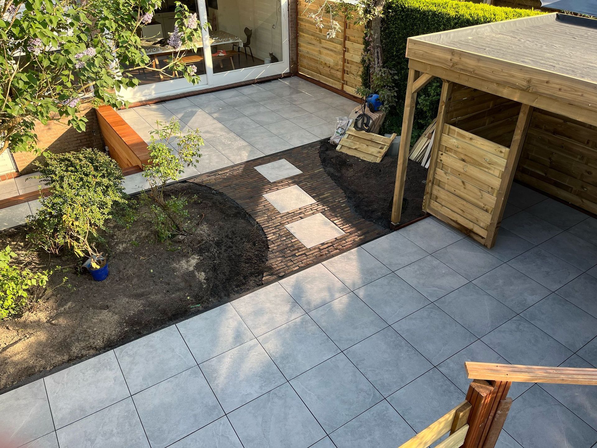 Terras met grijze tegels, een houten schuur, een pad met stapstenen en groen.