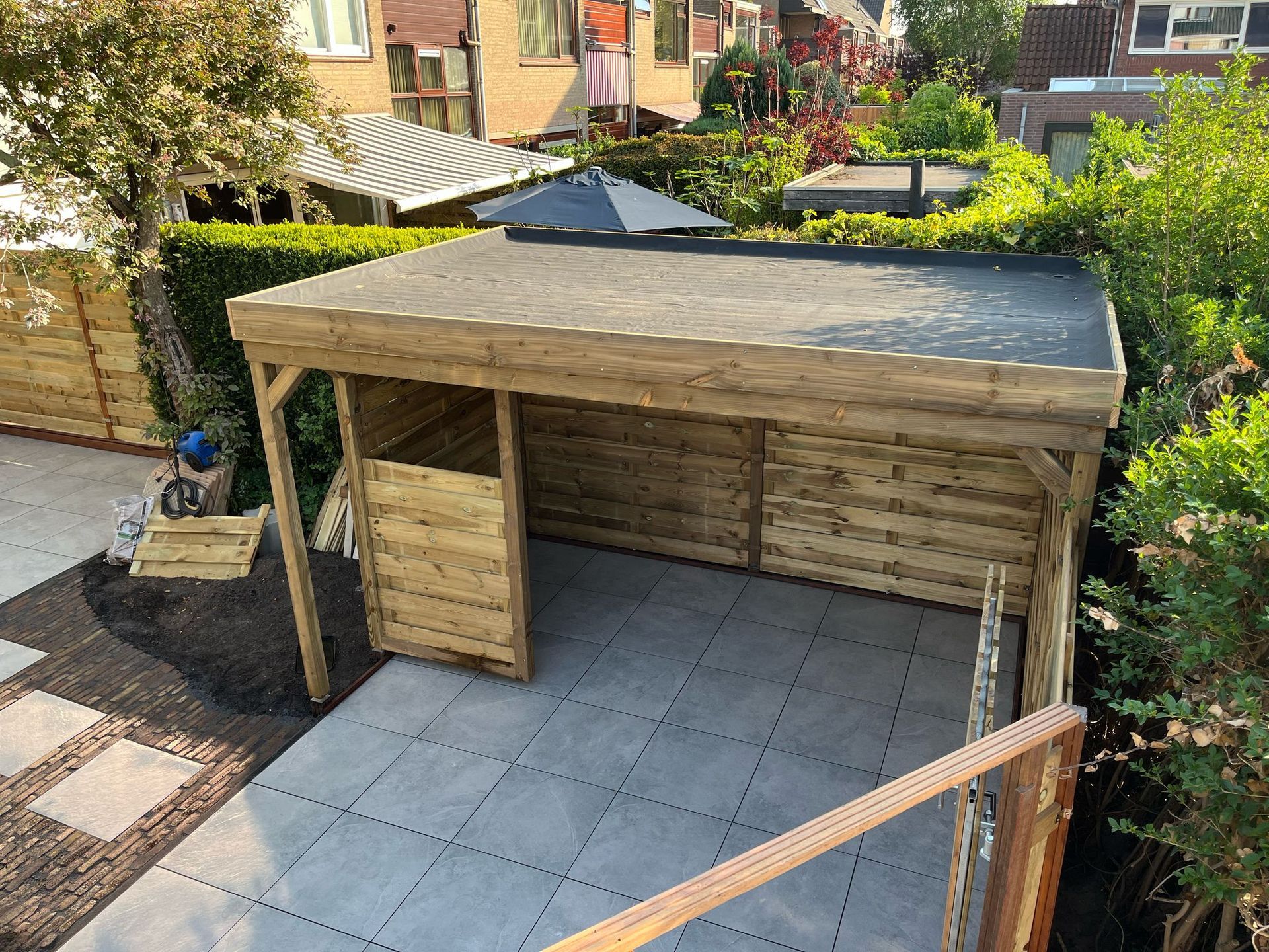 Houten schuur met plat, zwart dak in een achtertuin met een grijs bestraat terras.