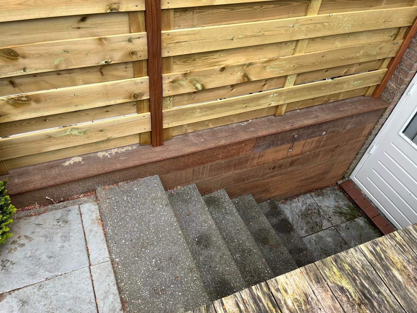 Een trap leidt vanaf een houten hek en een deuropening naar beneden. Grijze betonnen treden en een keermuur.