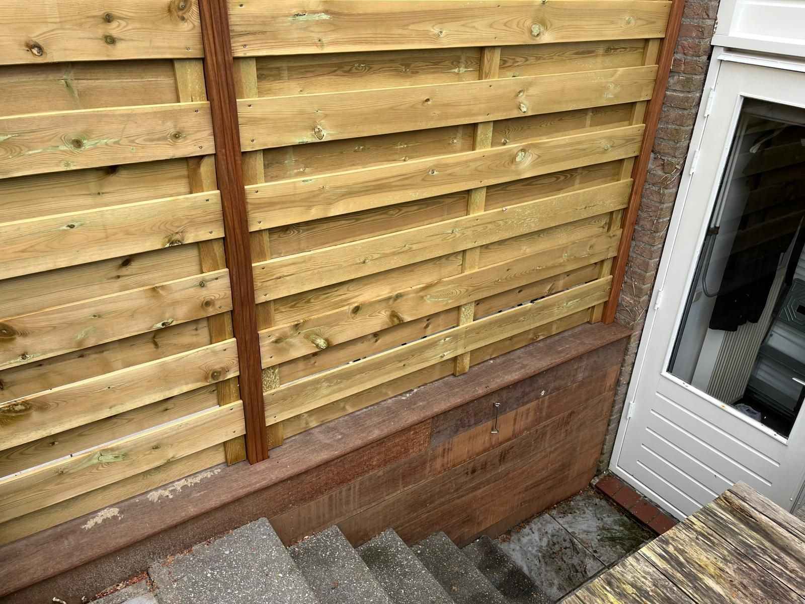 Houten schutting met horizontale planken naast een witte deur en betonnen trappen.