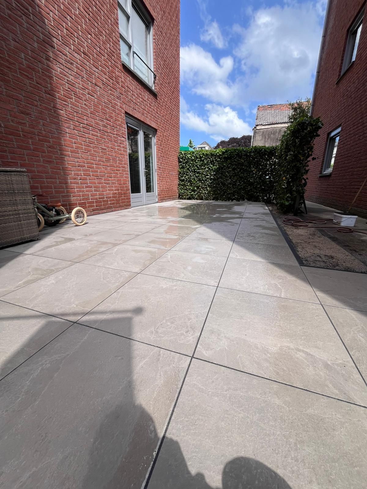 Bakstenen gebouwen flankeren een terras met grijze tegels. Op de achtergrond zijn een heg en een zonnige hemel te zien.