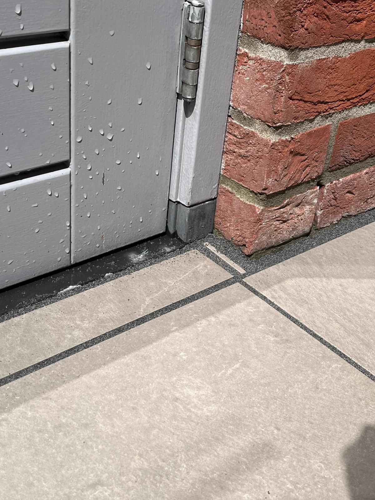 Close-up van een grijs poortscharnier bevestigd aan een bakstenen muur en een betonnen ondergrond met waterdruppels.