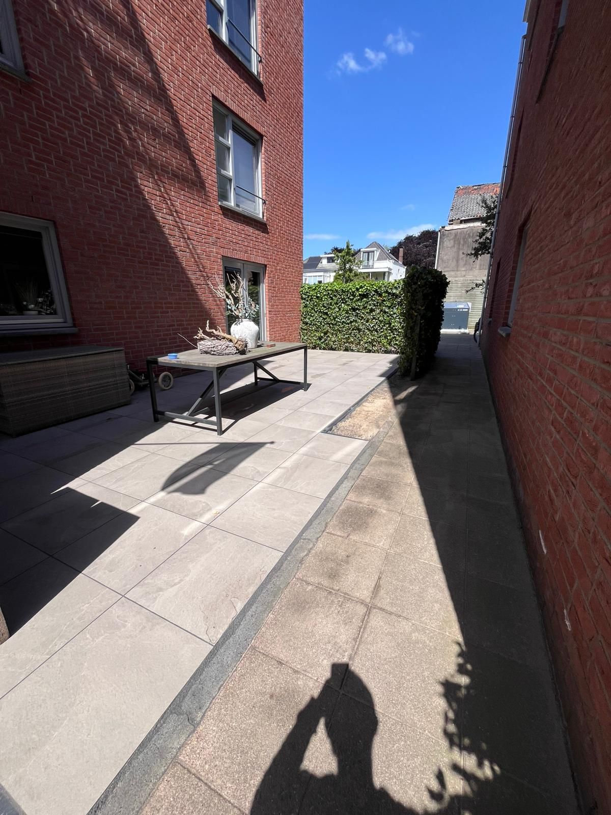 Bakstenen gebouwen flankeren een zonovergoten terras. Een tafel staat bij een heg, met een figuur in de schaduw die een foto maakt.