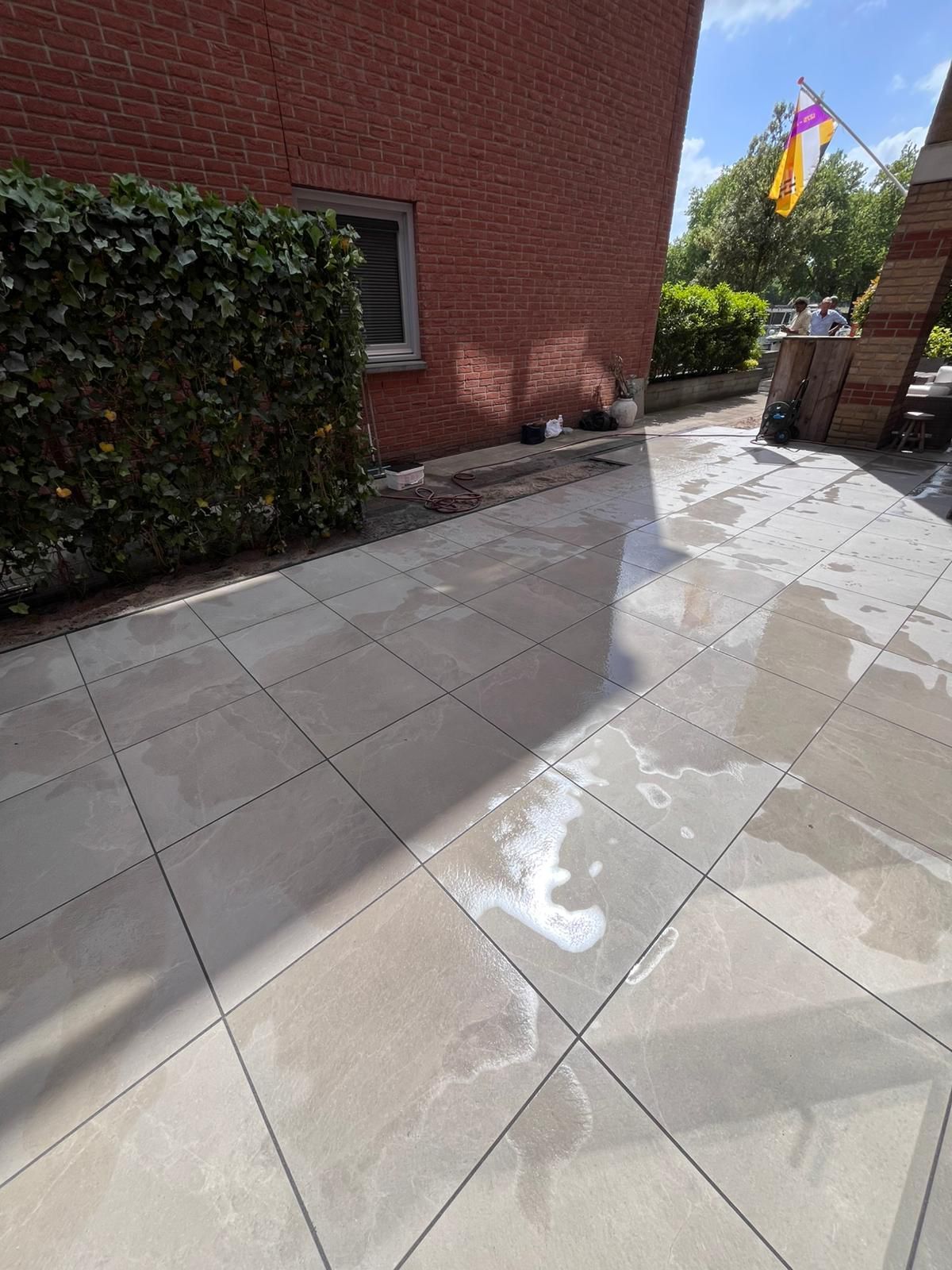 Een nat, lichtgekleurd stenen terras naast een rood bakstenen gebouw en een groene heg.