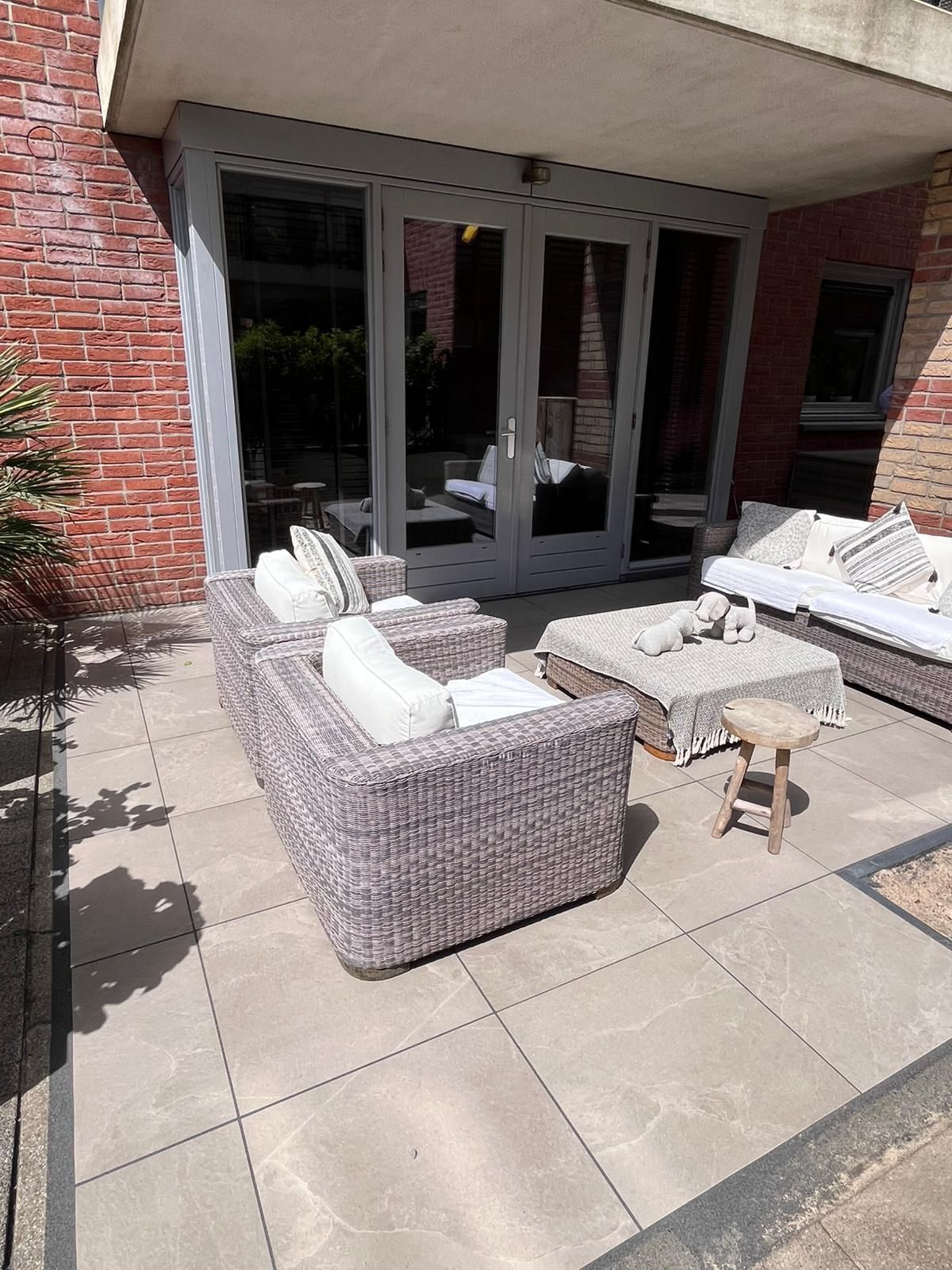 Terras met zitgedeelte, voorzien van rieten meubels en glazen deuren. Op de achtergrond een gebouw van rode baksteen.