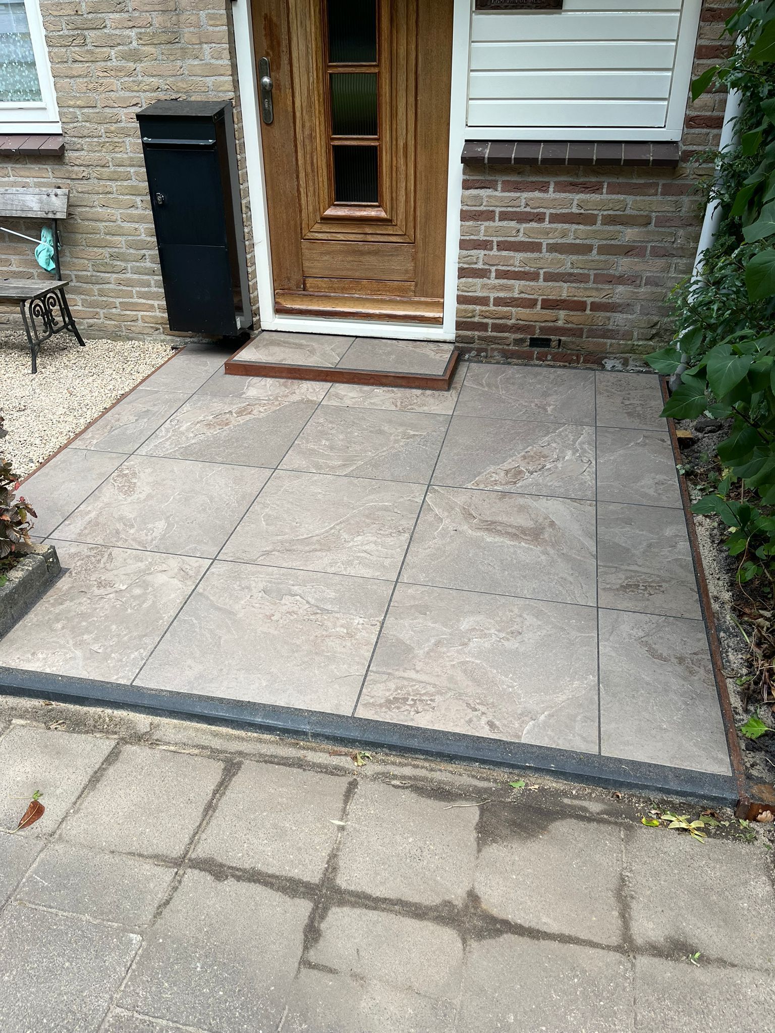 Stenen terras voor een bakstenen gebouw. Een houten deur, een zwarte brievenbus en groen zijn zichtbaar.