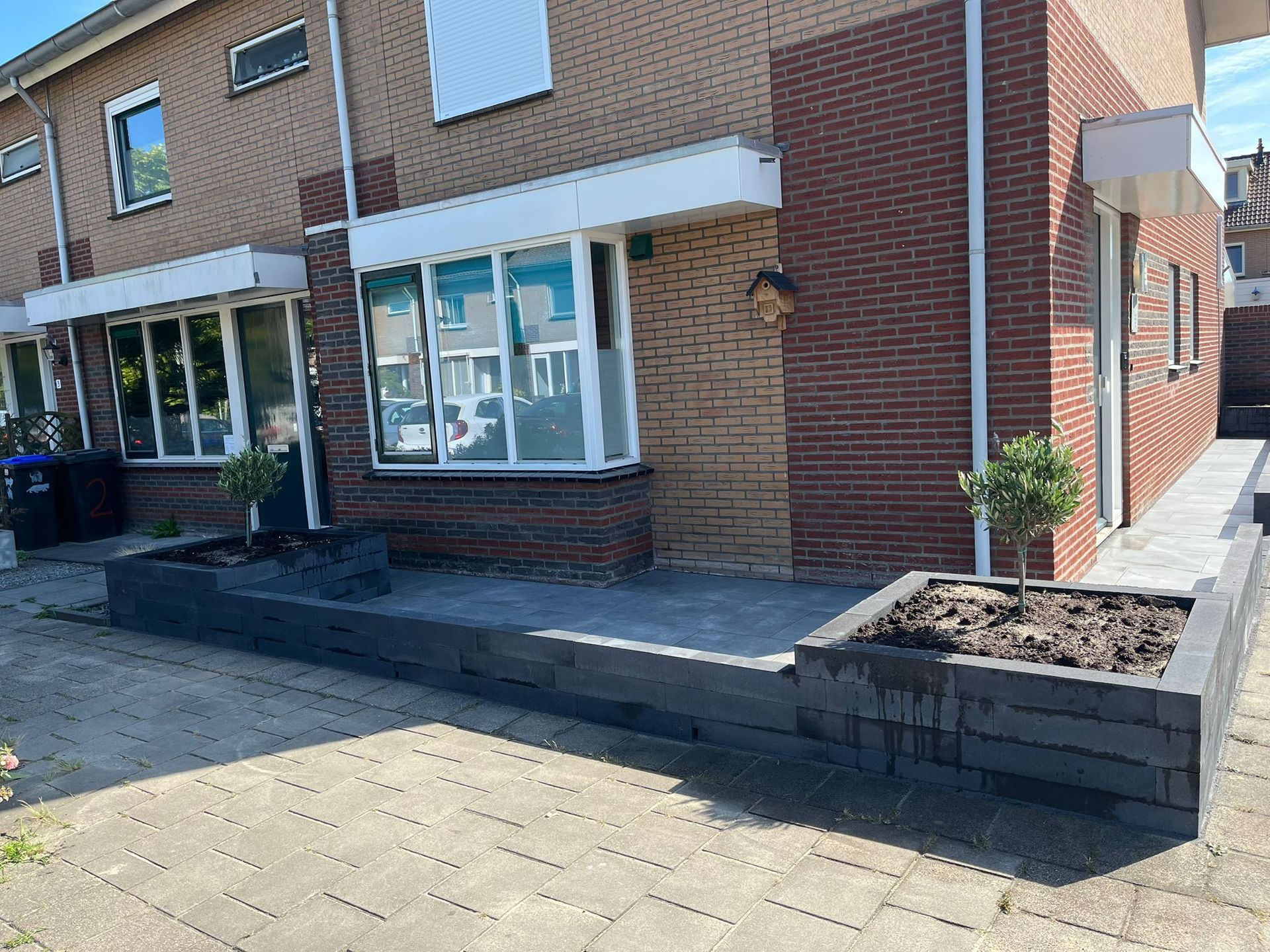 Een bakstenen gebouw met een betonnen terras en een donkere plantenbak ervoor. In de plantenbak staan ​​twee kleine boompjes.