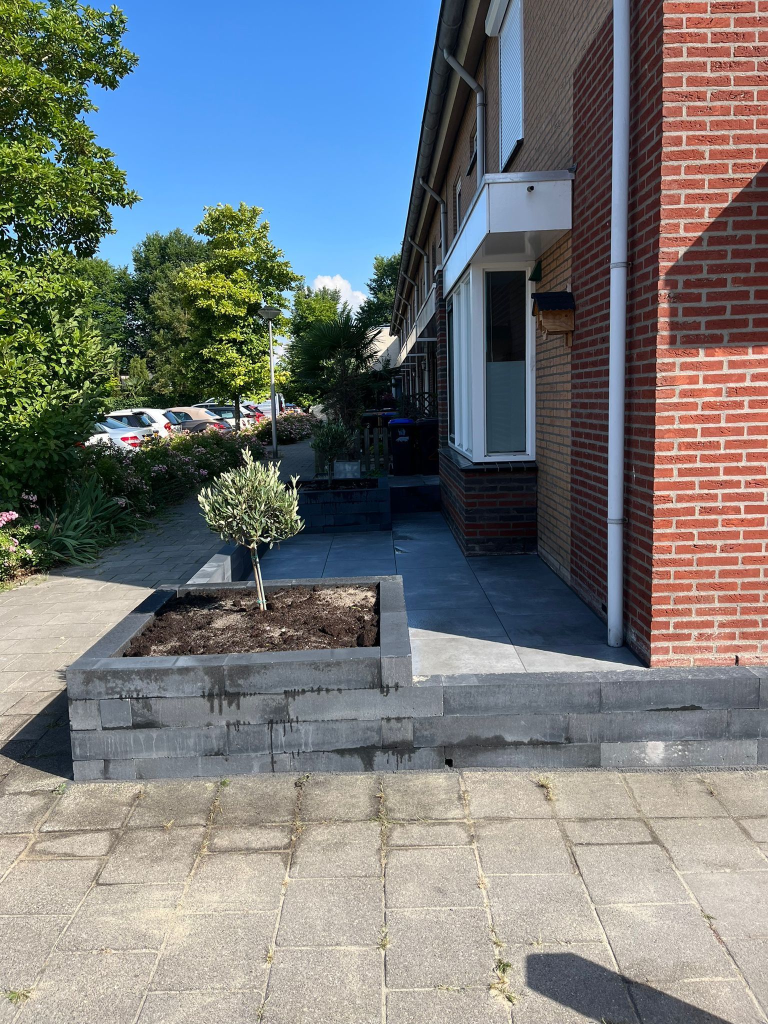De buitenkant van een huis met een bakstenen gevel en een donkergrijze stenen plantenbak met een kleine boom.