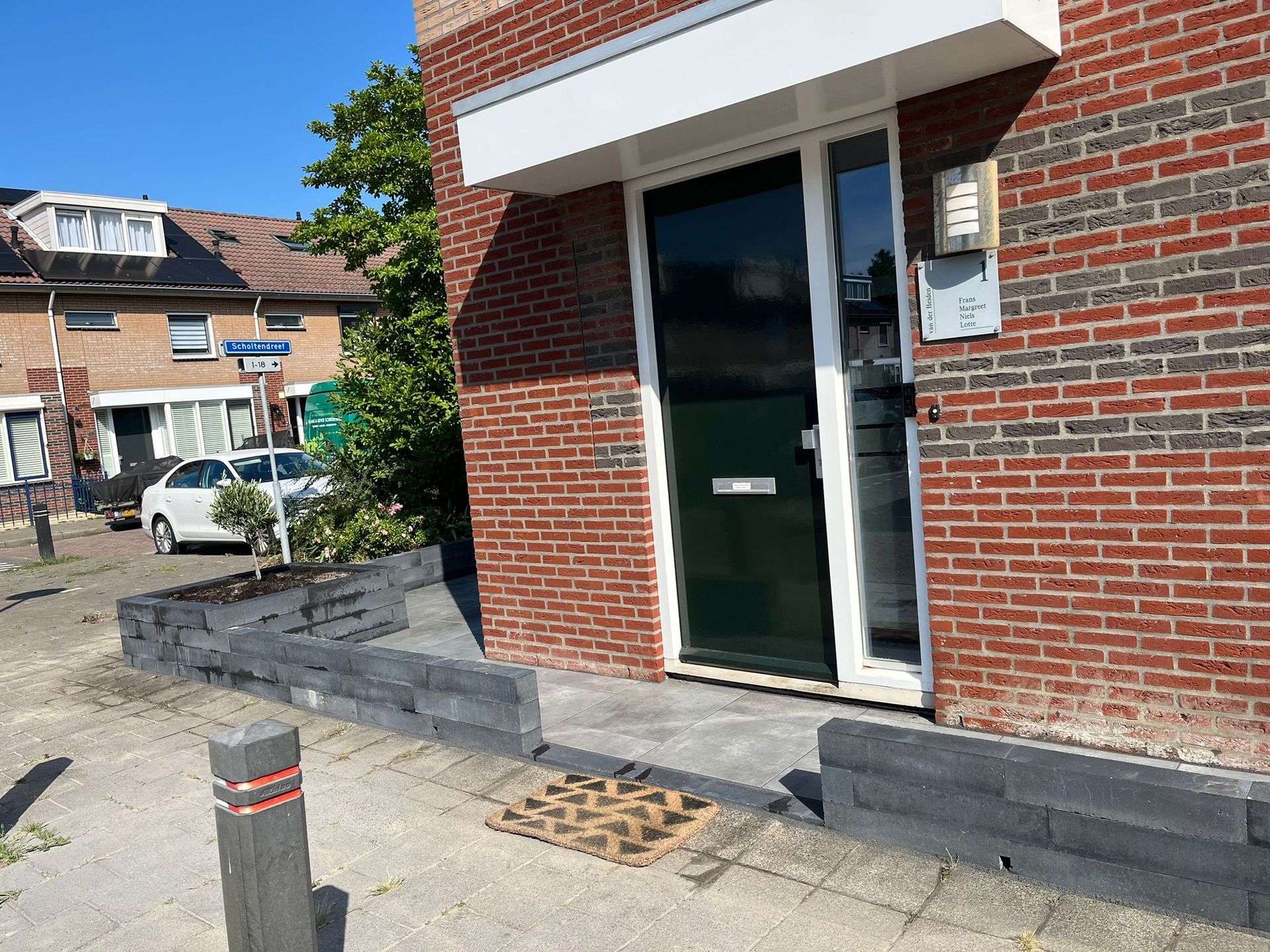 Ingang van een bakstenen gebouw met een donkere deur, een kleine grijze keermuur en een straatbeeld op een zonnige dag.