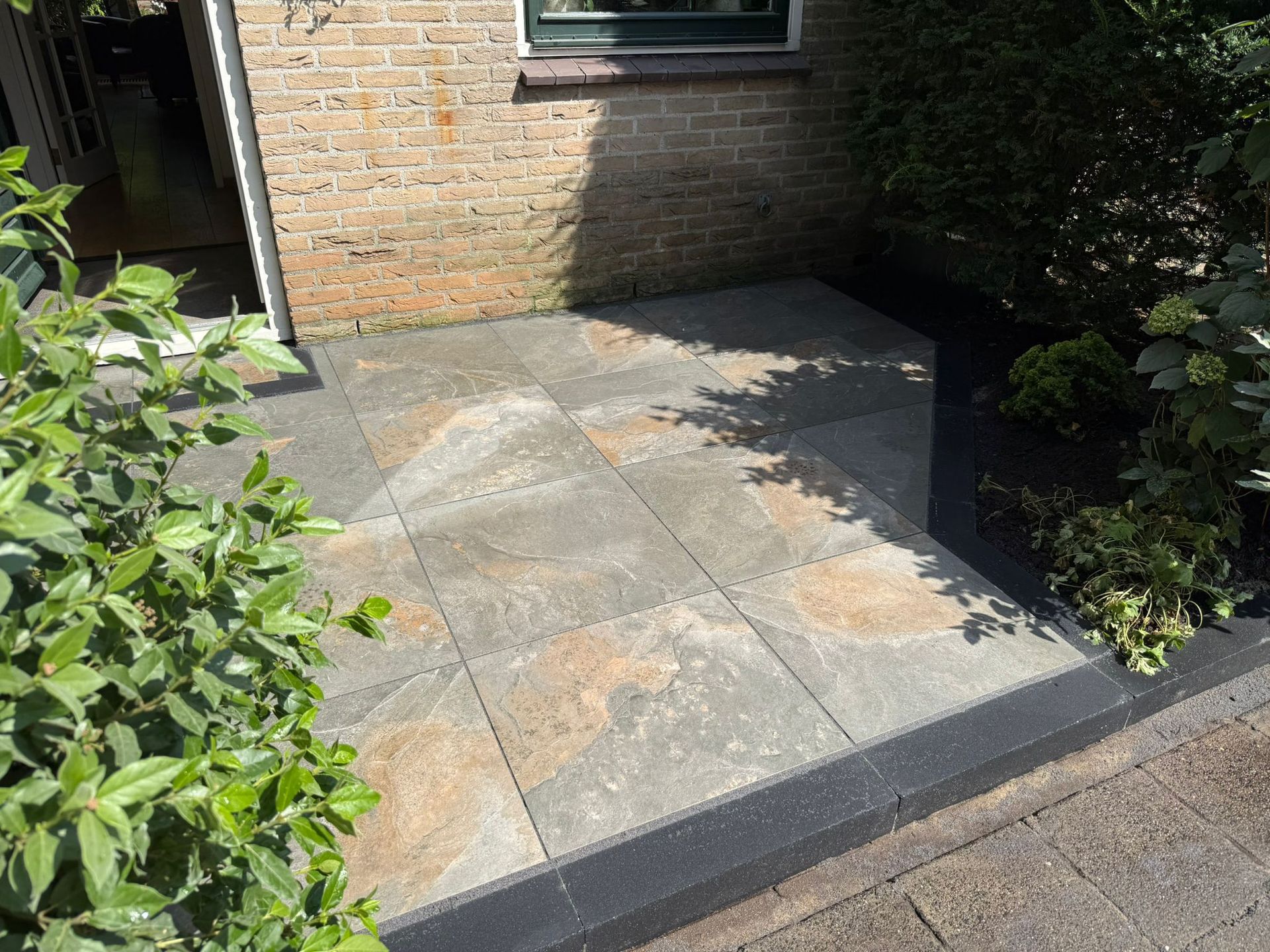 Terras met stenen bestrating naast een bakstenen gebouw en groen.