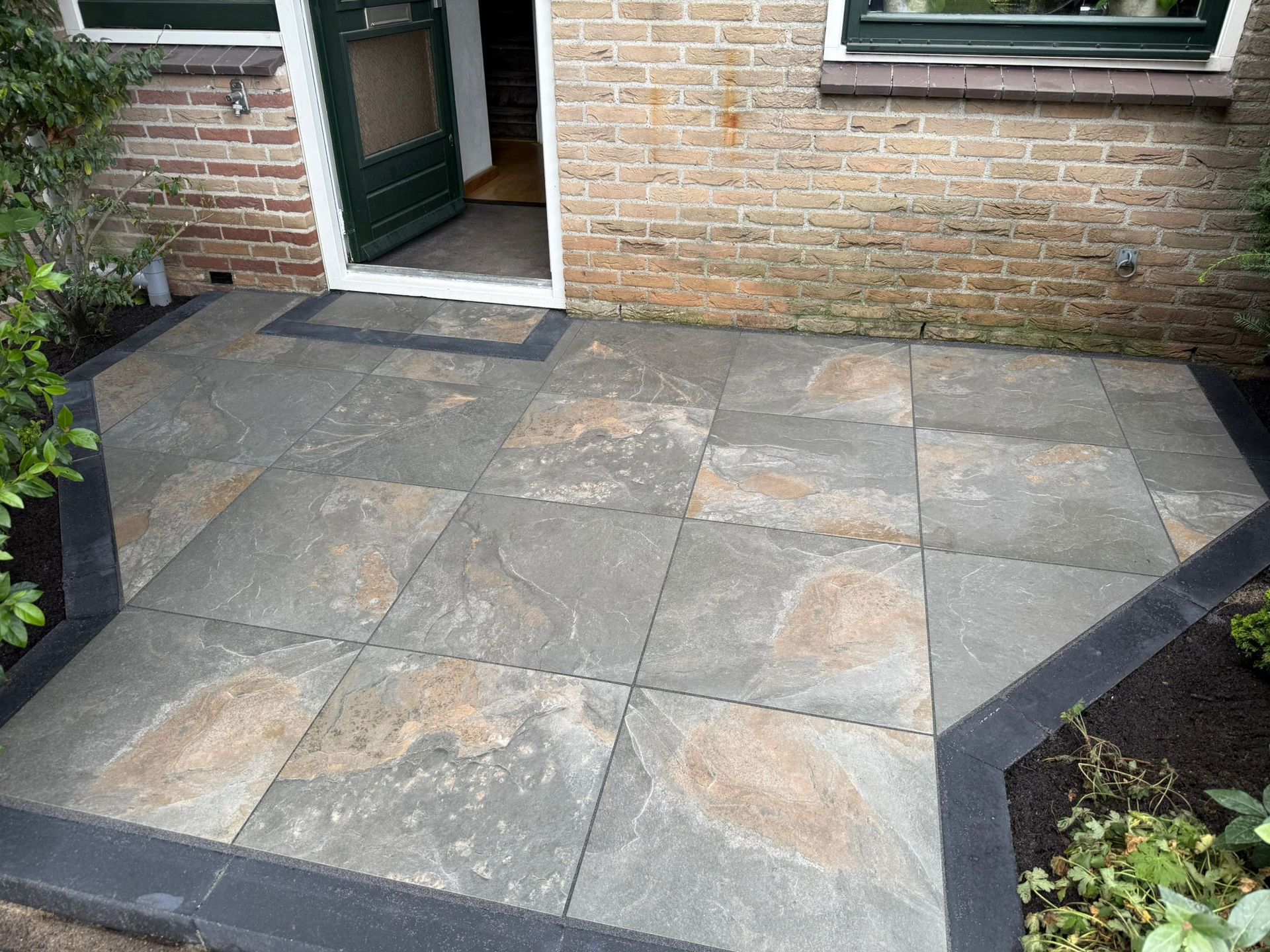 Terras met grijze plavuizen en een donkere rand naast een bakstenen gebouw met een groene deur.