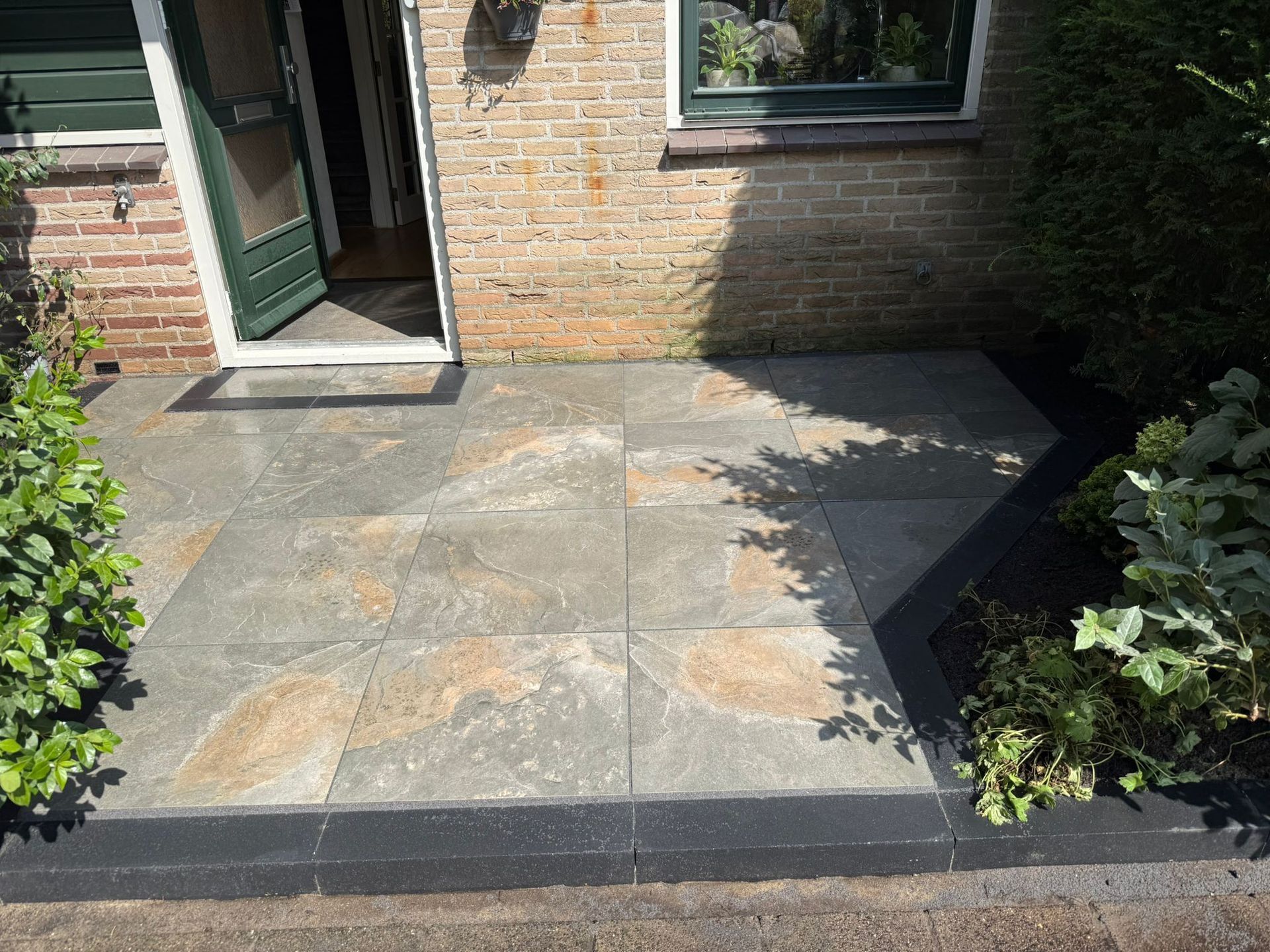Entree met grijze stenen tegels, bakstenen muur, groene deur en omringende beplanting.