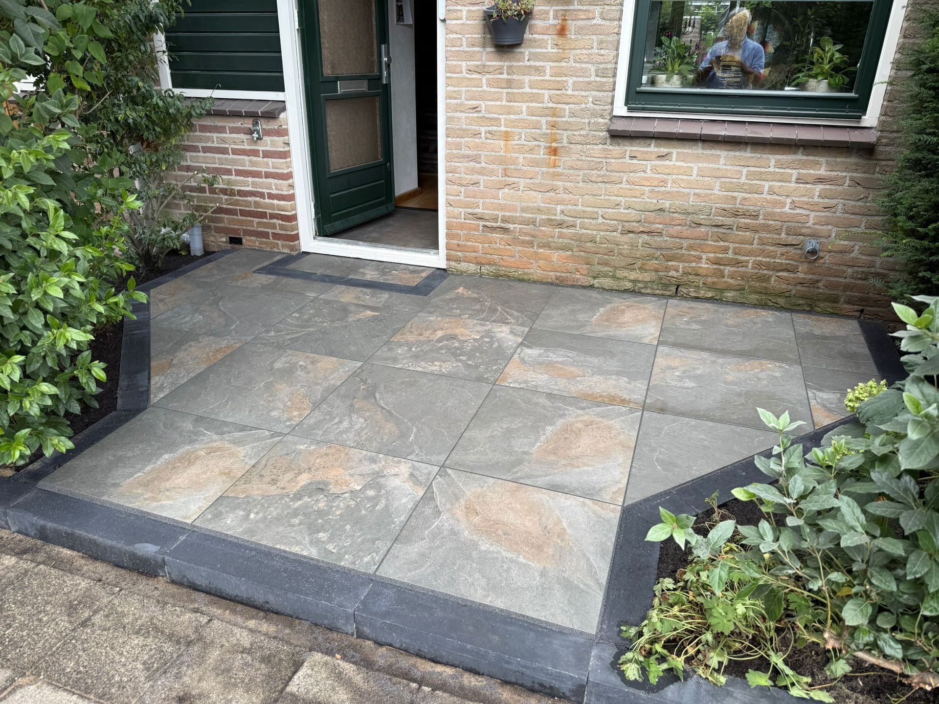 Terras met grijze natuurstenen tegels, omzoomd door een zwarte rand, voor een bakstenen gebouw met een open deur.