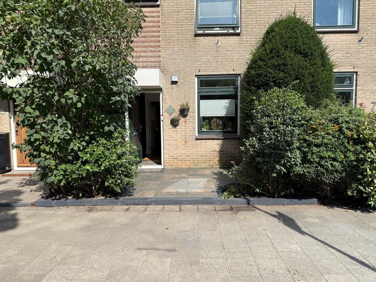 Een bakstenen gebouw met een open deur, geflankeerd door struiken en bomen aan een geplaveid pad.