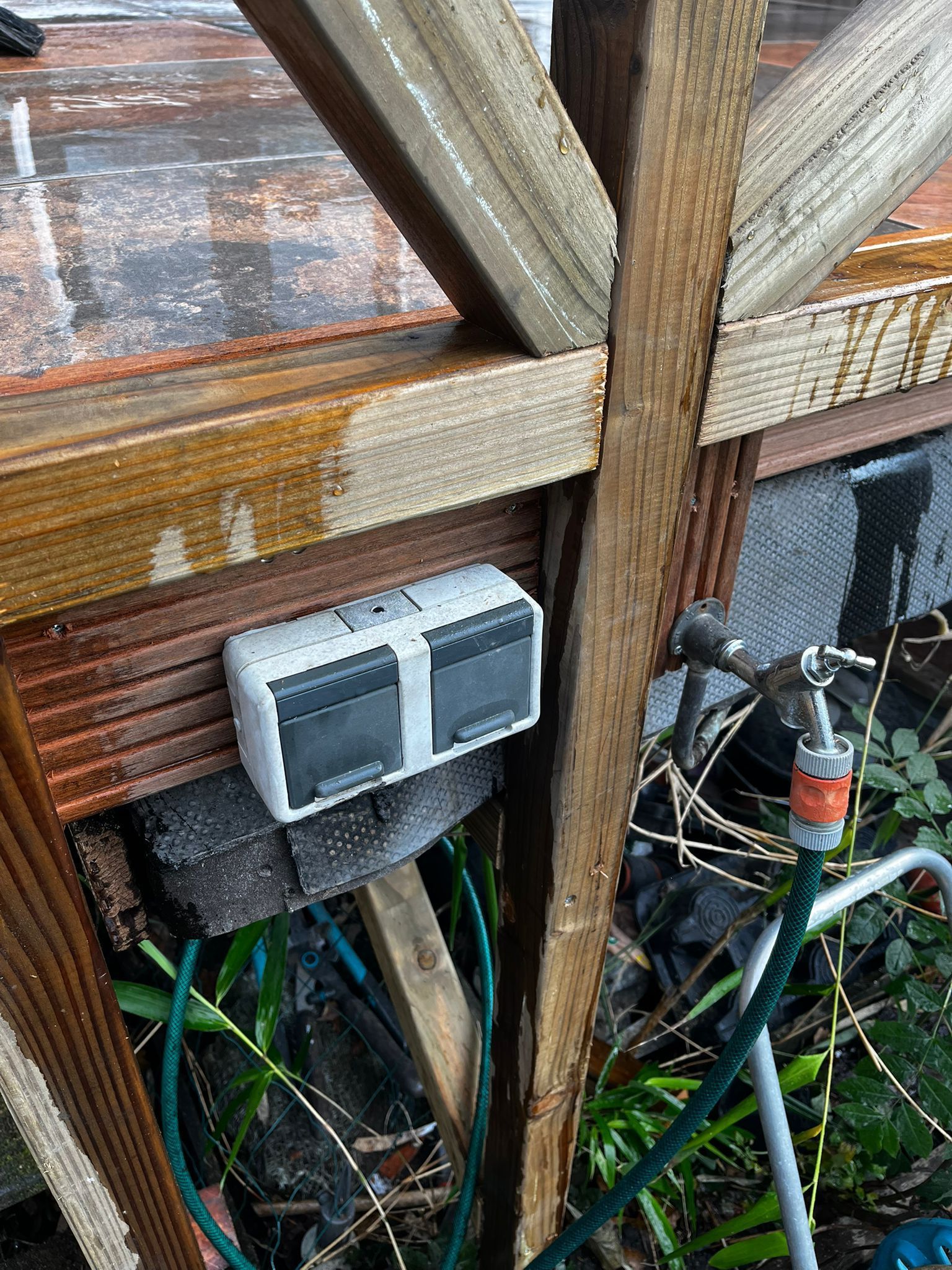 Een timerkastje voor de waterregeling is bevestigd aan een houten constructie vlakbij de buitenkraan met slang.