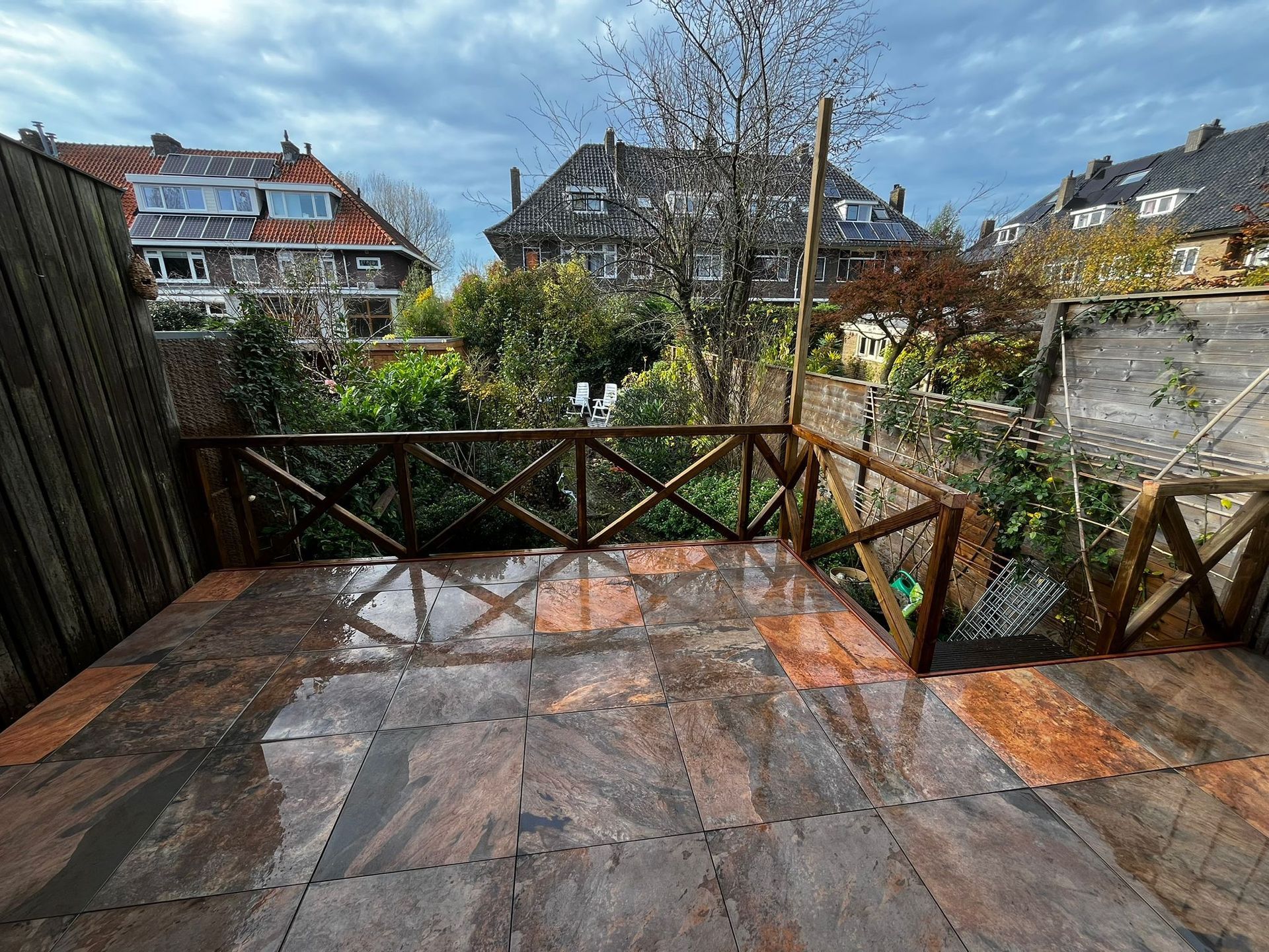 Terras met natuurstenen tegels en houten balustrade, met uitzicht op een tuin met huizen op de achtergrond onder een bewolkte hemel.