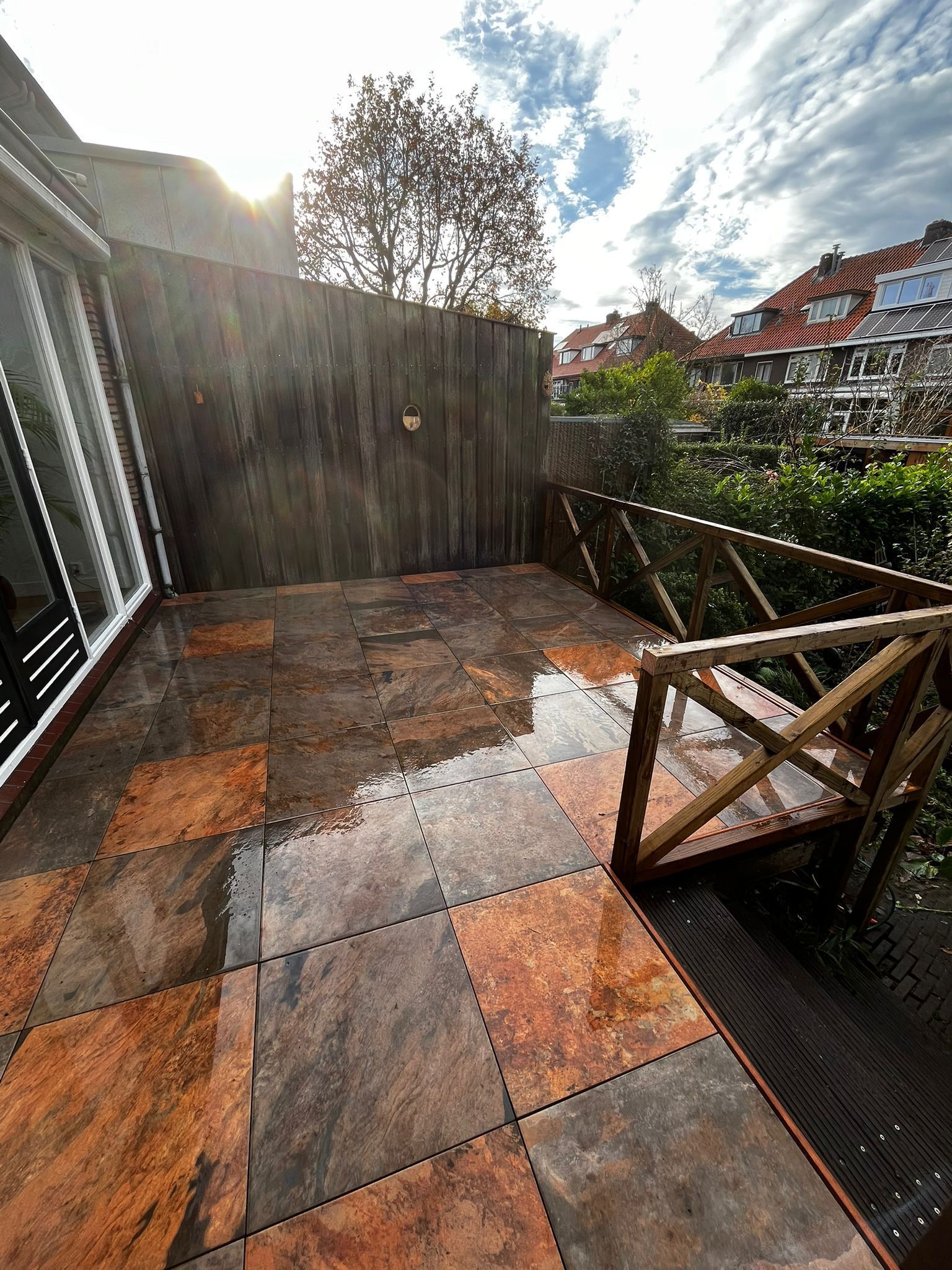 Terras met natte, patroonrijke tegels, houten balustrade en een rustieke houten muur. Zonnige dag.