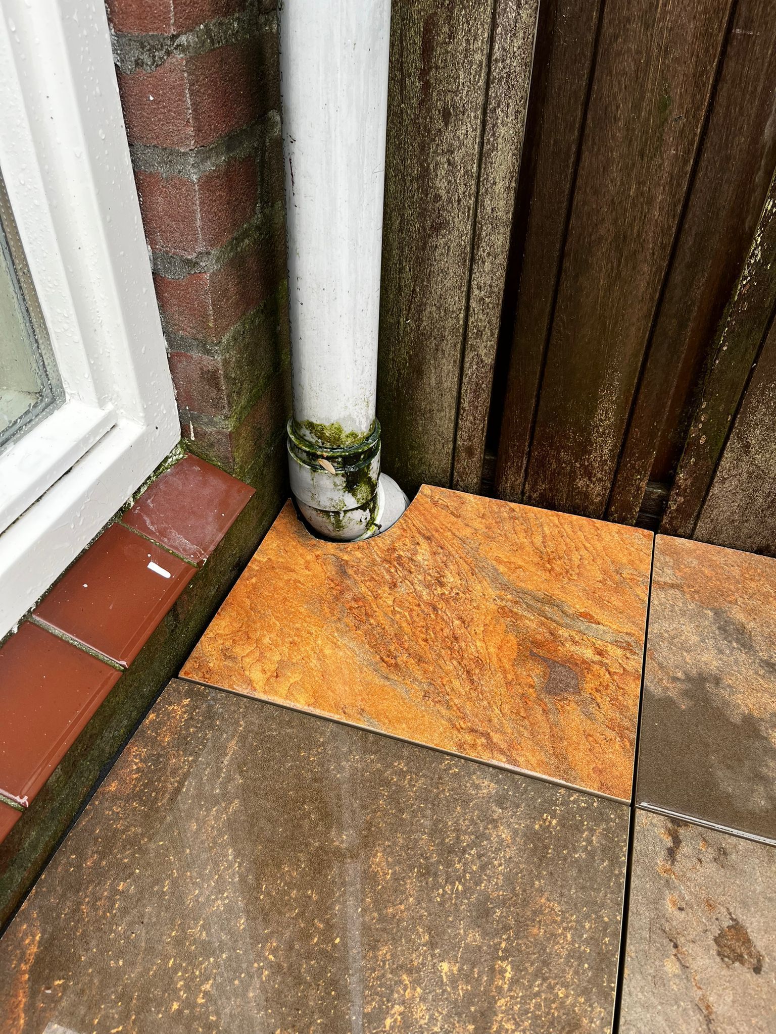 Regenpijp die afwatert op roestbruine terrastegels naast een bakstenen muur en houten schutting.