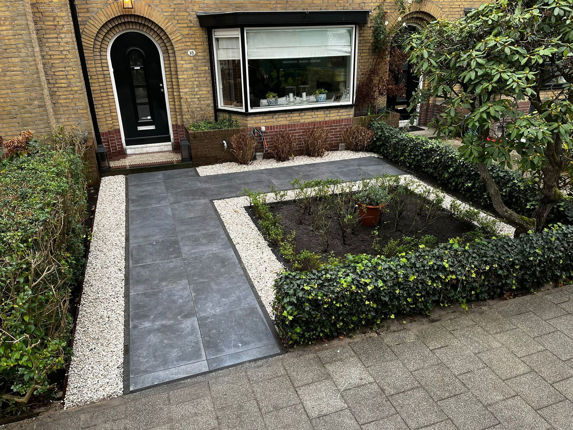 Voortuin met een bestraat pad, bloemperk en een deur naar een bakstenen huis.