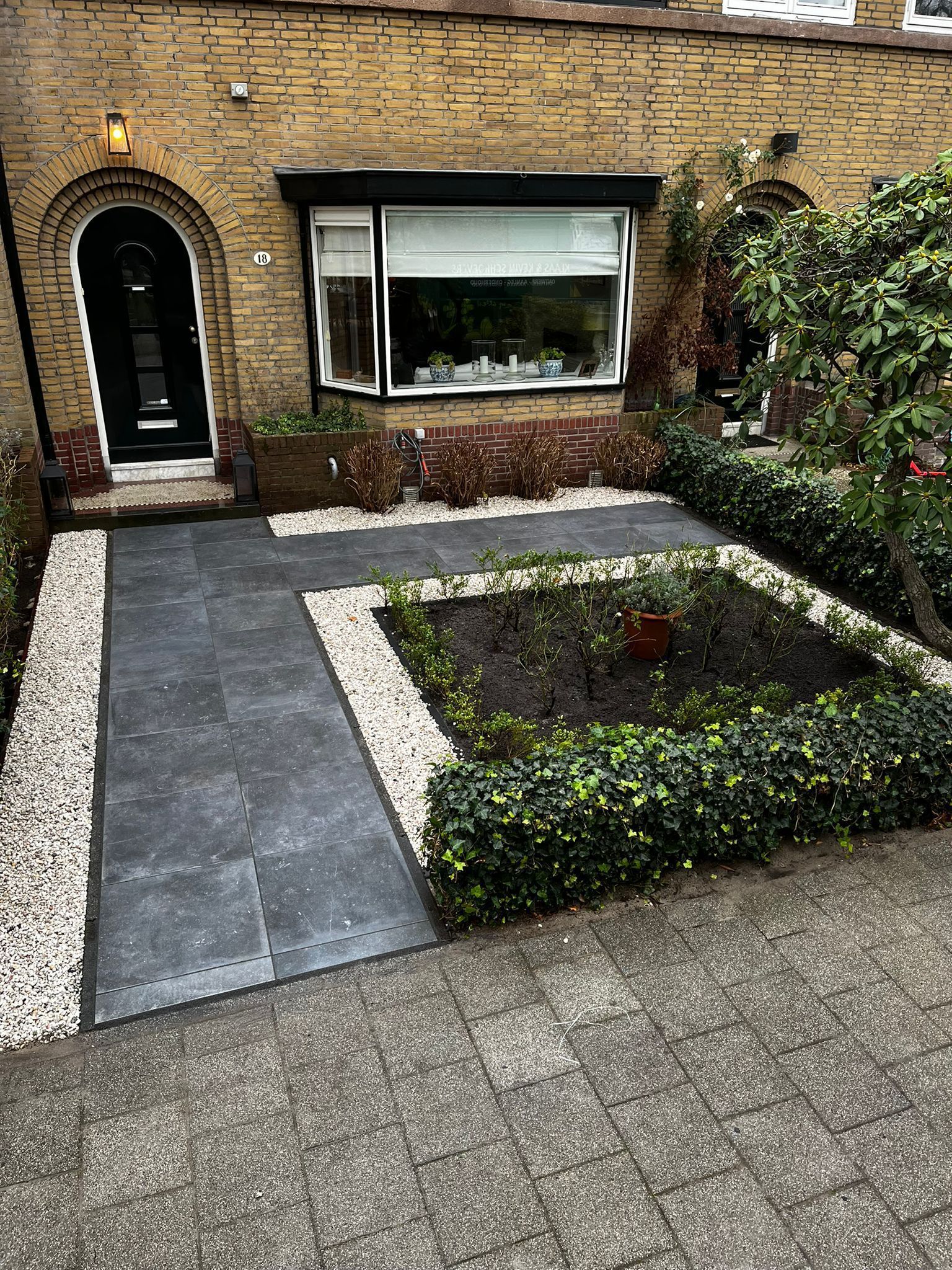 Voortuin met grijze bestrating, witte stenen en een kleine tuin omzoomd door groene hagen.