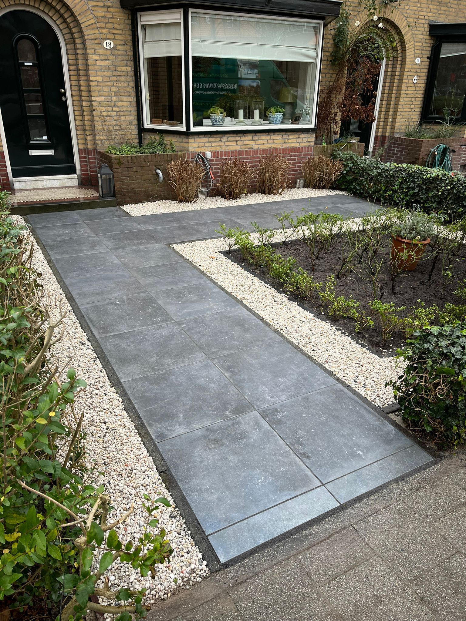 Een betonnen pad leidt naar de ingang van een gebouw. ​​De rand van het pad is bedekt met grind. Aan weerszijden bevinden zich beplanting en struiken.