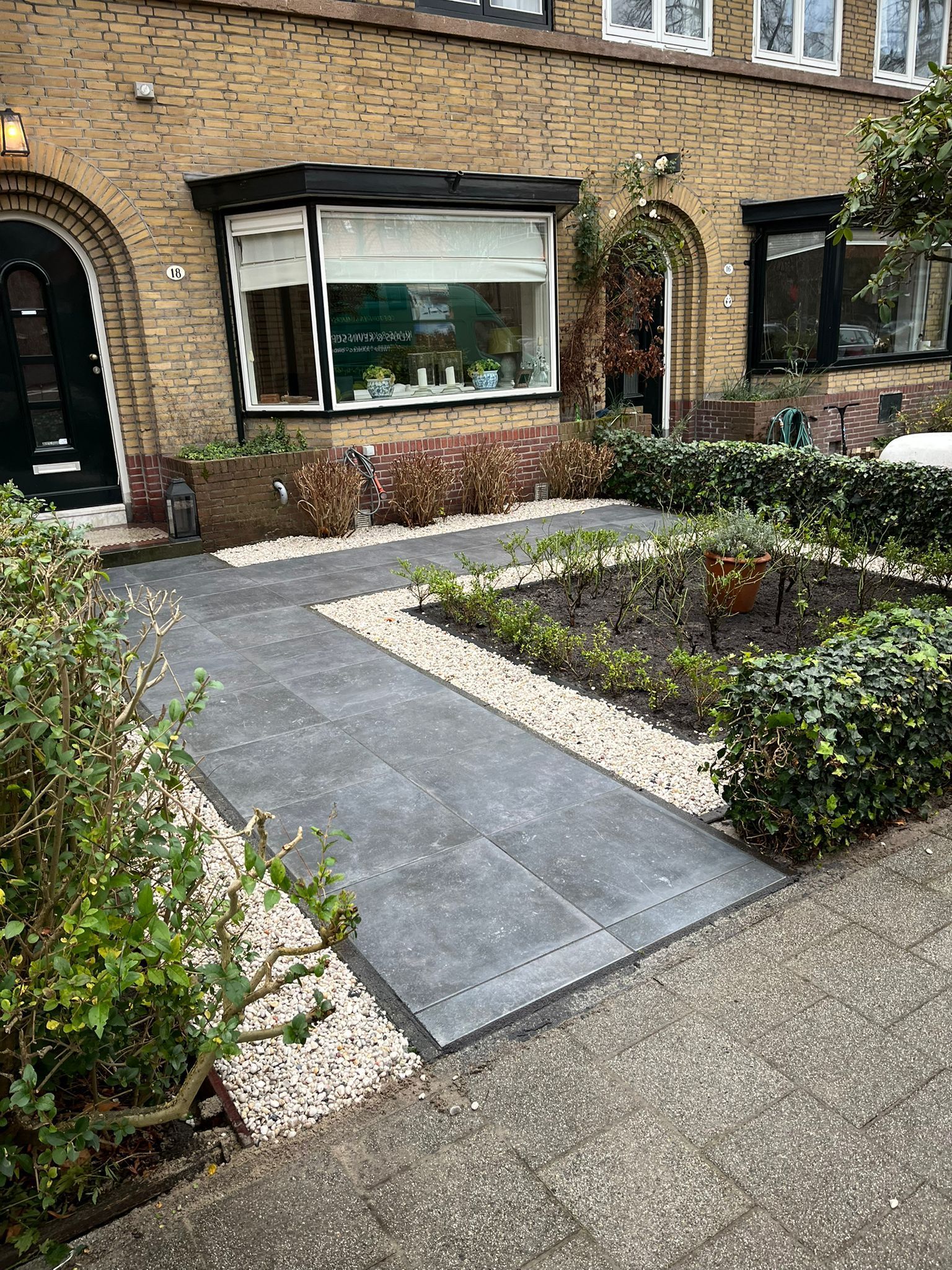 Pad naar een huis met grind en planten. Bakstenen buitenkant, ramen met zwarte kozijnen.