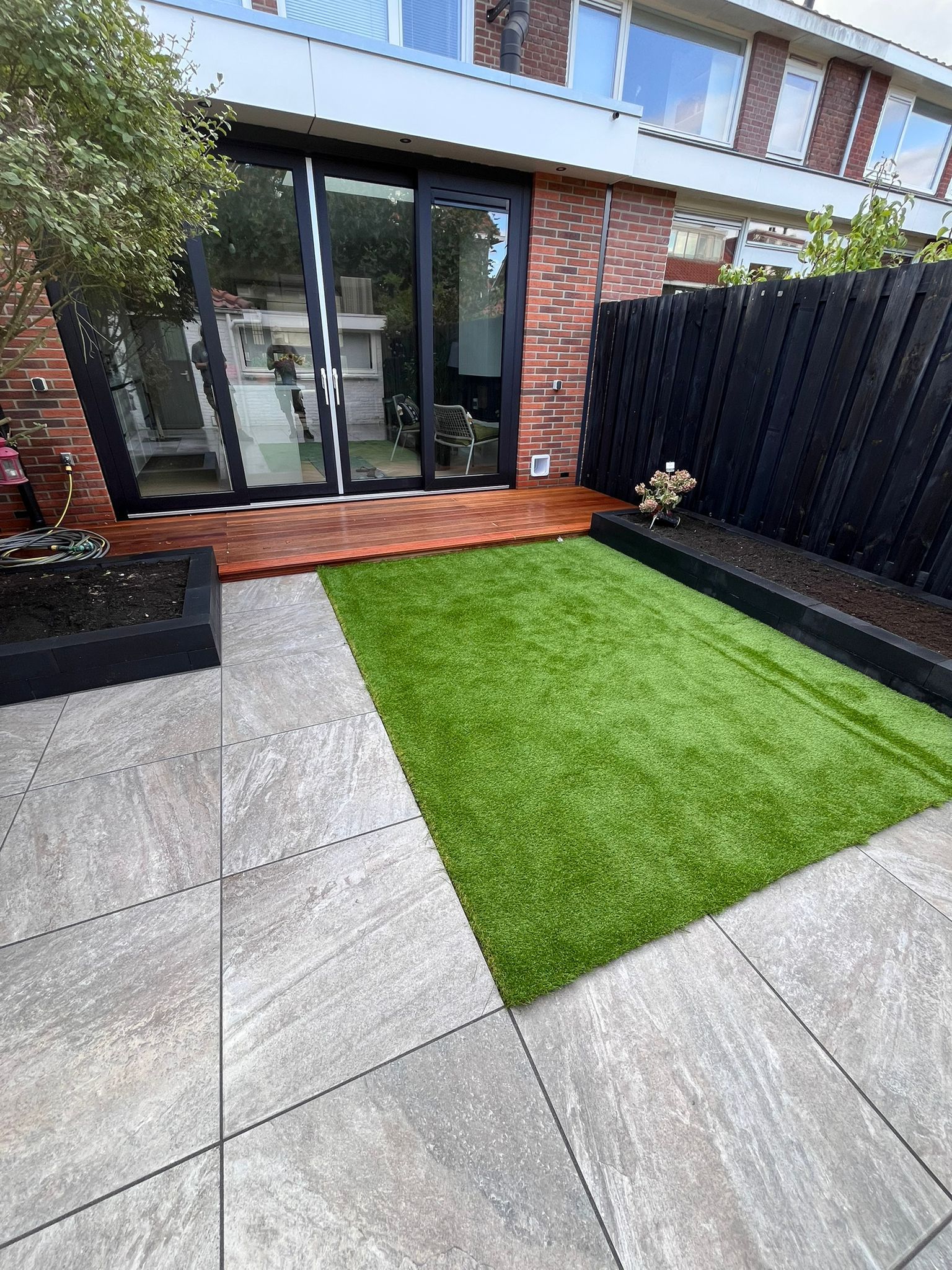 Een kleine achtertuin met een strook kunstgras, een houten terras en een zwart hek.