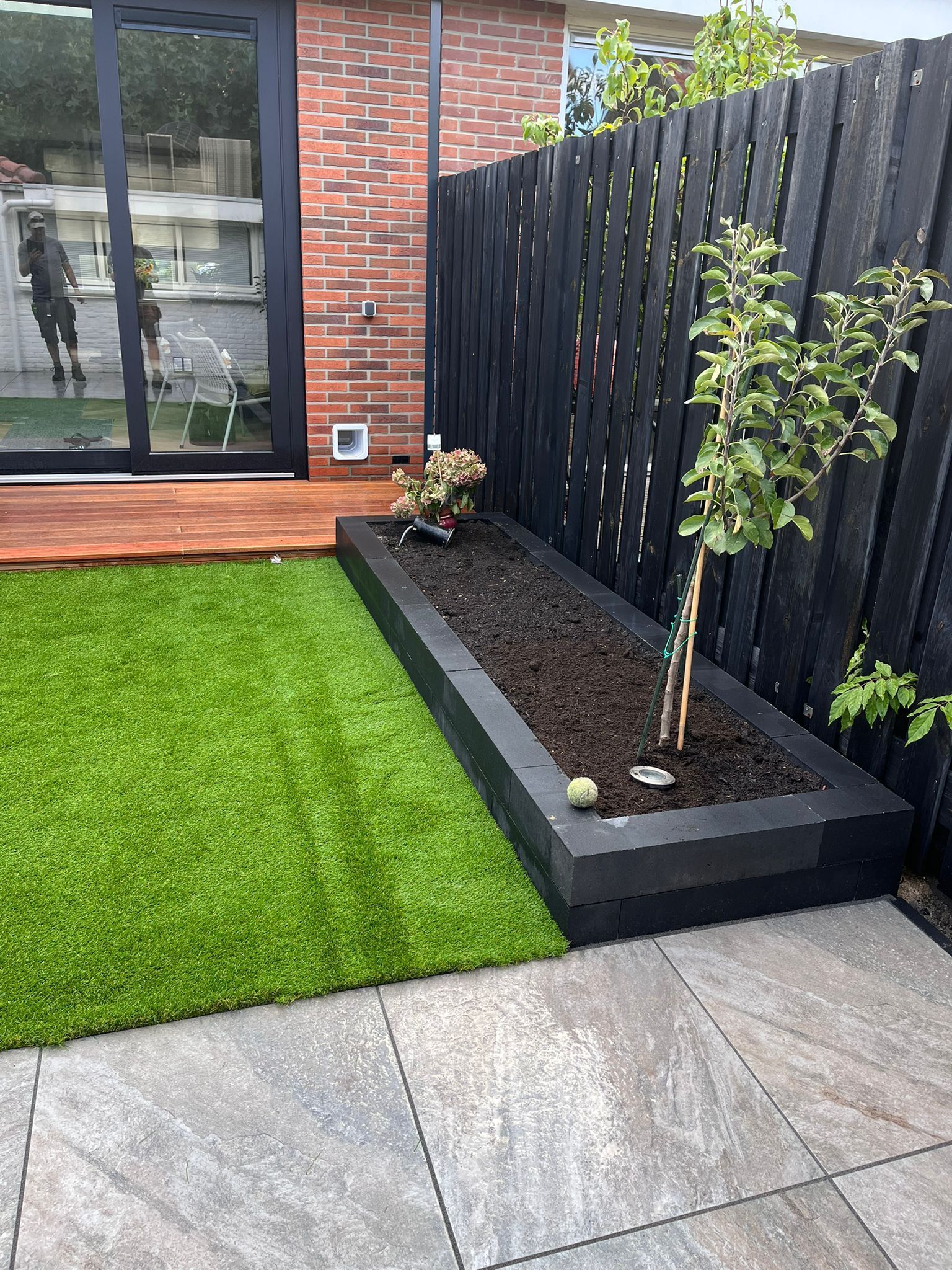 Een groen gazon en een stenen terras grenzen aan een zwarte plantenbak met een kleine boom vlakbij een zwart hek.