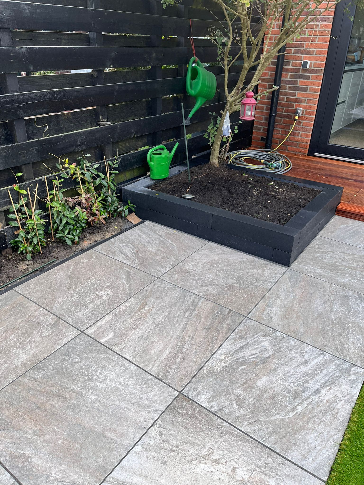 Terras in de achtertuin met grijze tegels, een zwarte plantenbak en een zwart hek.
