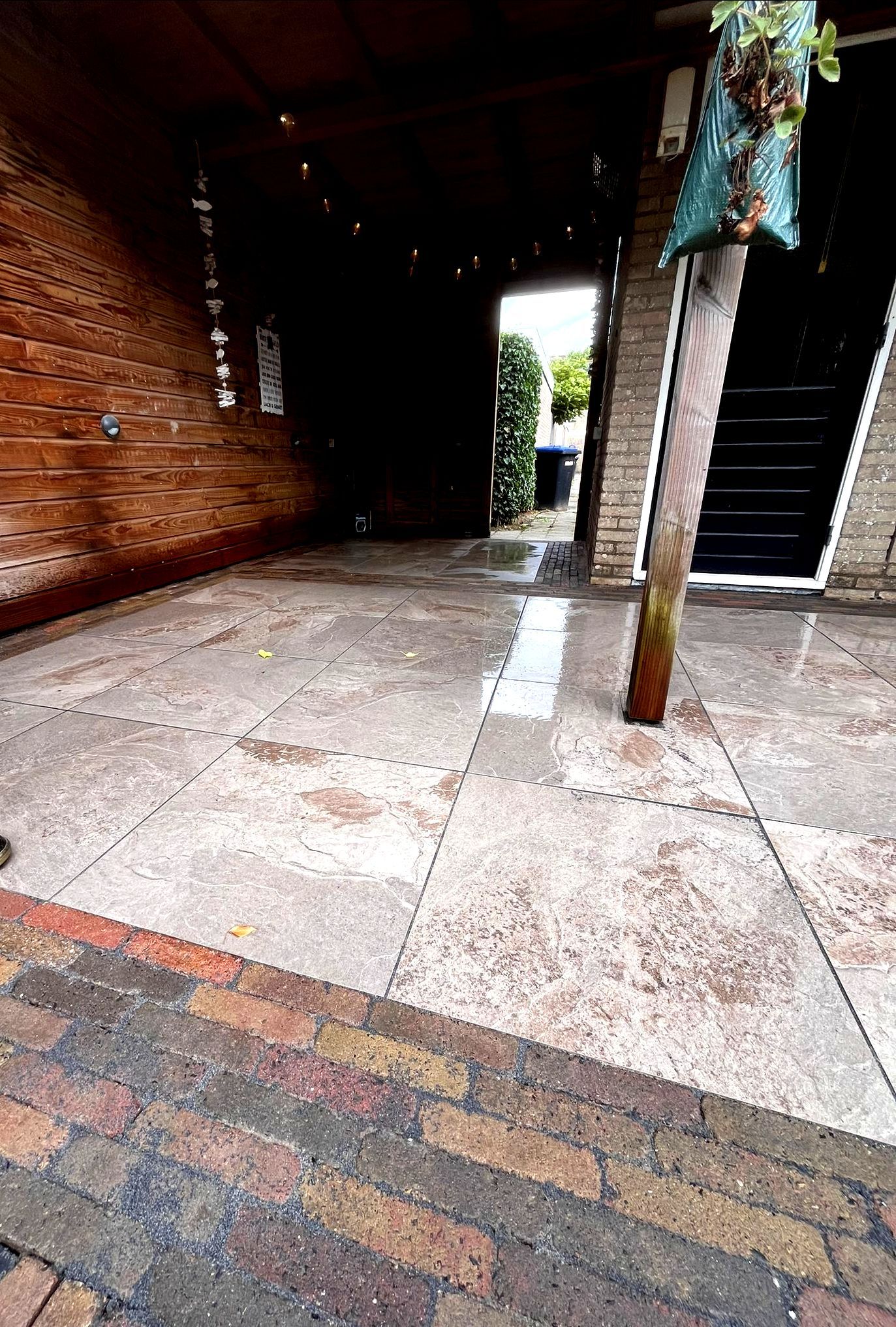 Een betonnen terras met een bakstenen rand, dat leidt naar een donkere gang met een deur en een bakstenen muur.