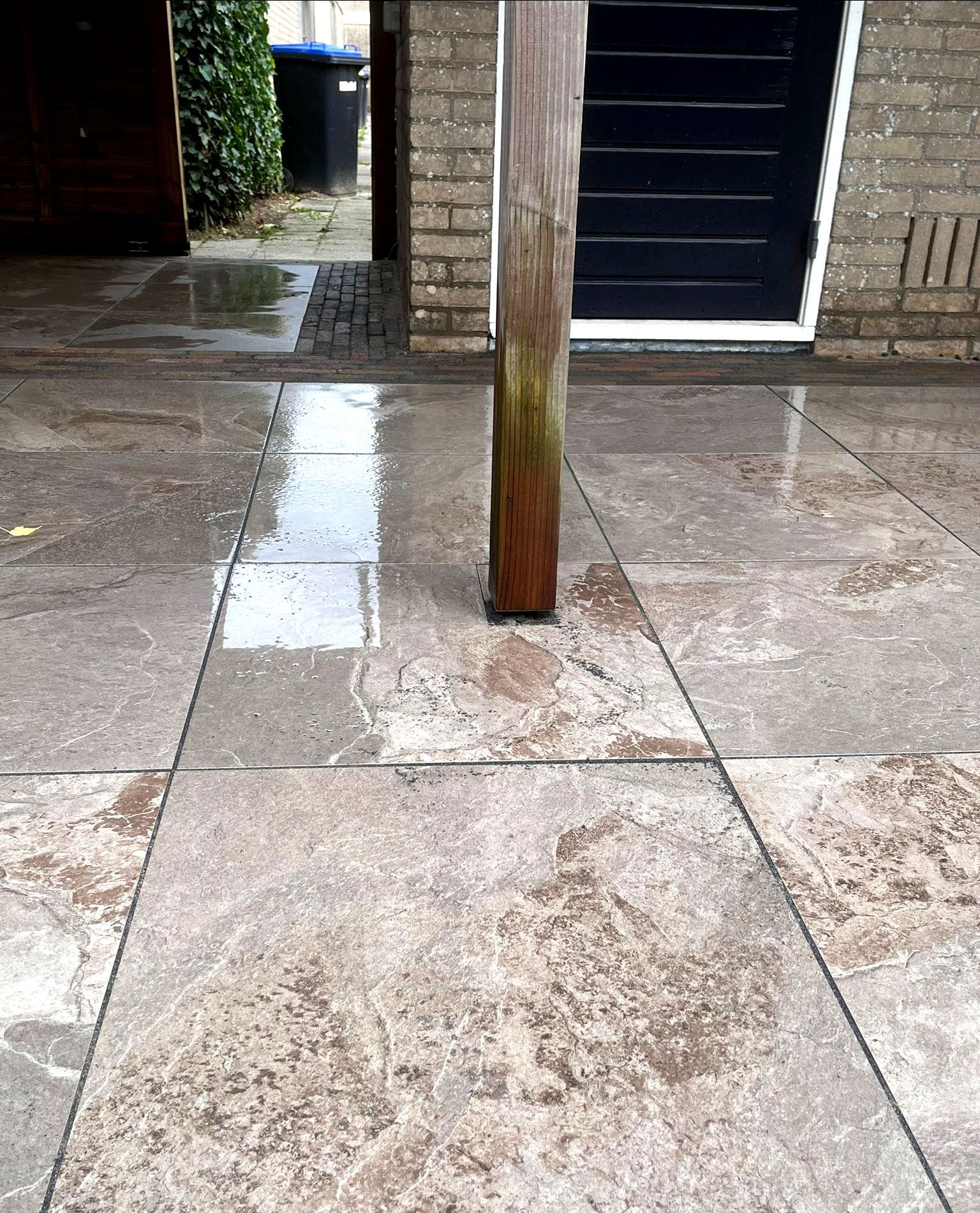 Een natte, betegelde vloer met een houten paal in het midden. Op de achtergrond zijn een ingang en bakstenen muren te zien.