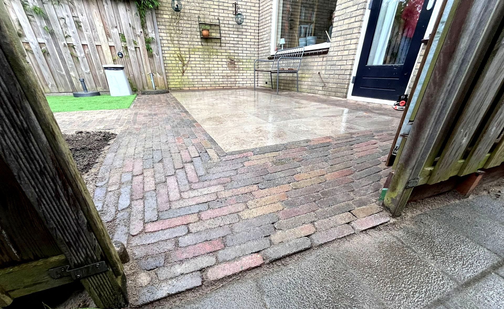 Een bakstenen pad leidt naar een terras, omgeven door een hek en een bakstenen gebouw.