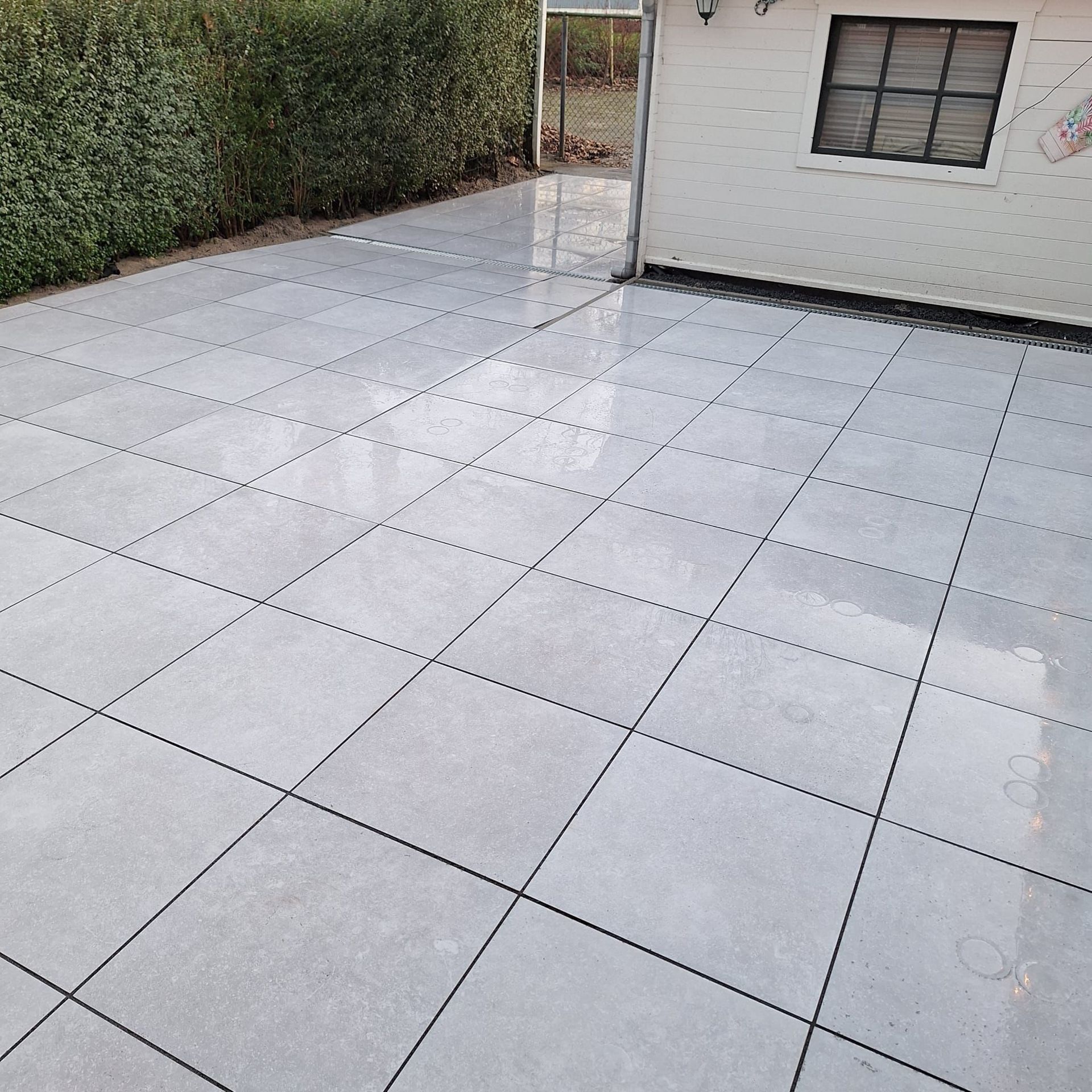 Een terras met grijze tegels naast een wit gebouw en een groene heg.