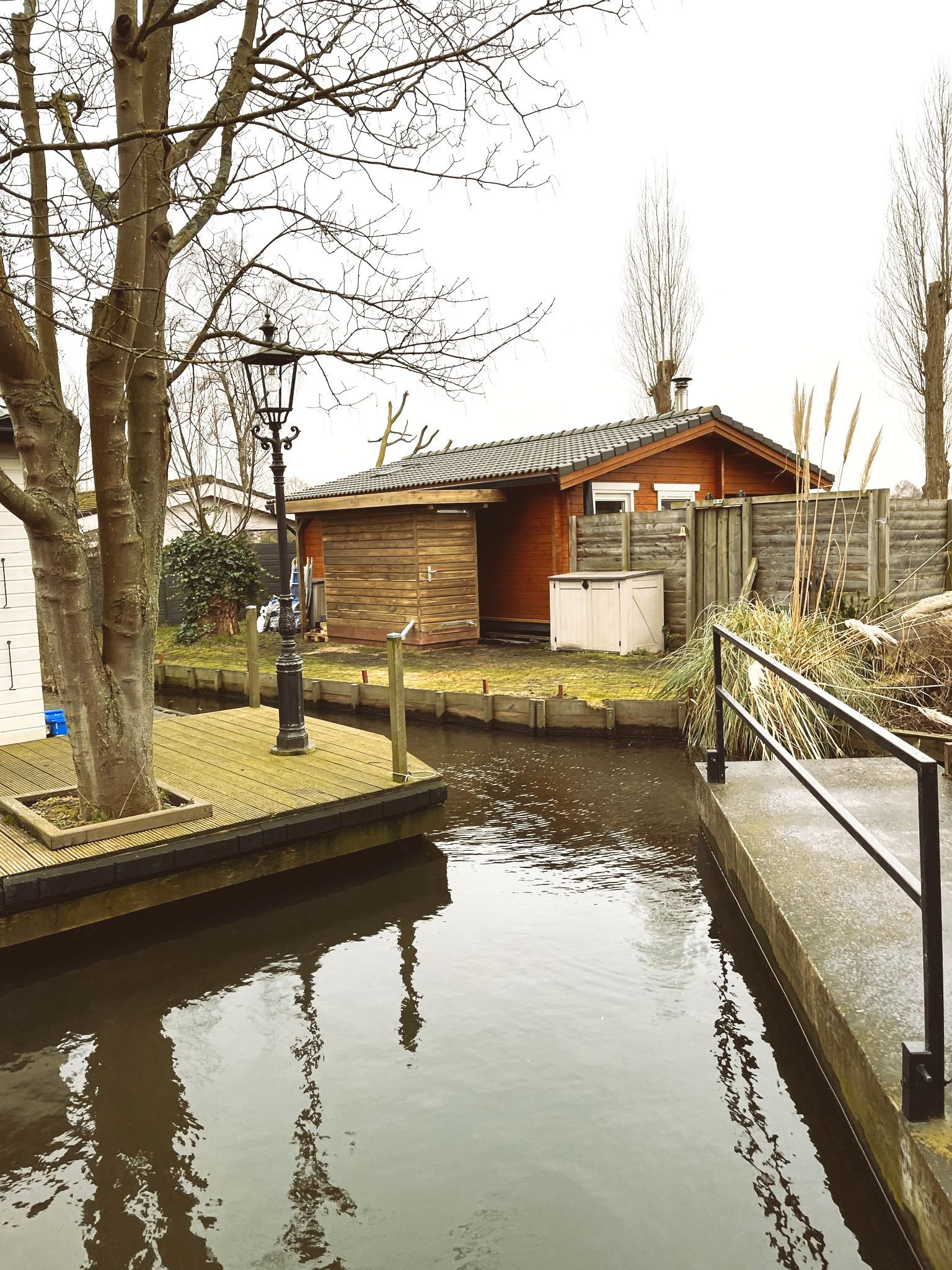 Een kanaal met houten gebouwen, bomen en een lantaarnpaal op de oever.
