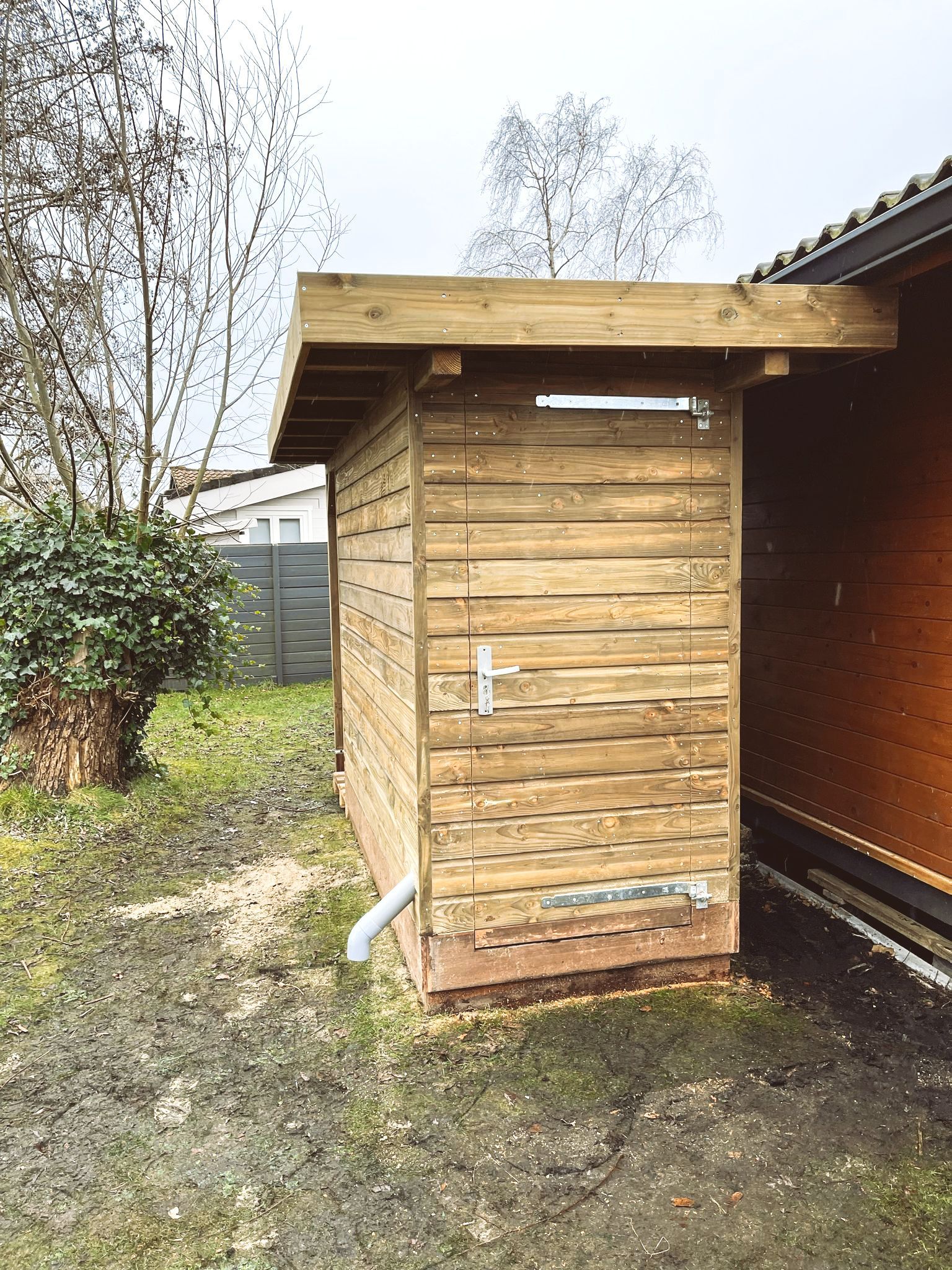 Houten schuur met deur en dak, gelegen naast een houten constructie, op een erf.