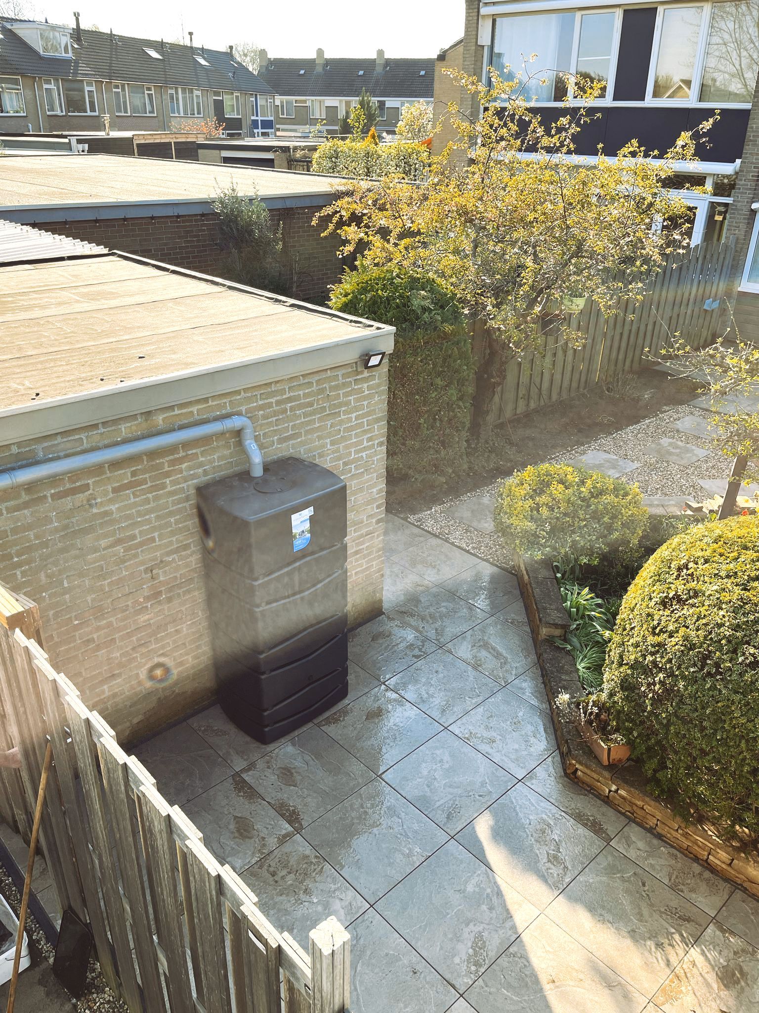 Bakstenen gebouw met regenwateropvangtank; terras, schutting en boom.