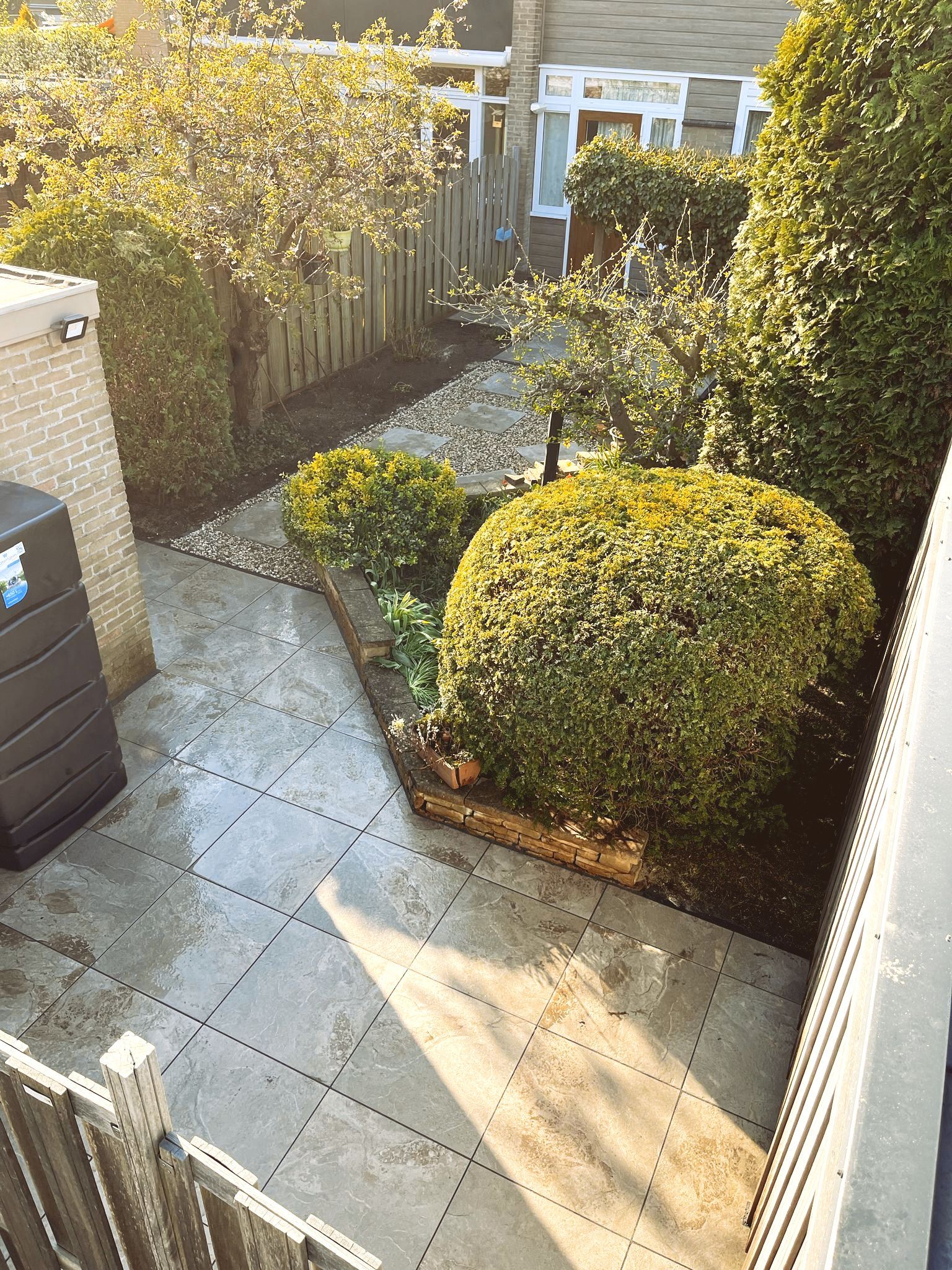 Een achtertuin met een terras, bloembedden en een schutting. De zon schijnt op het groen.