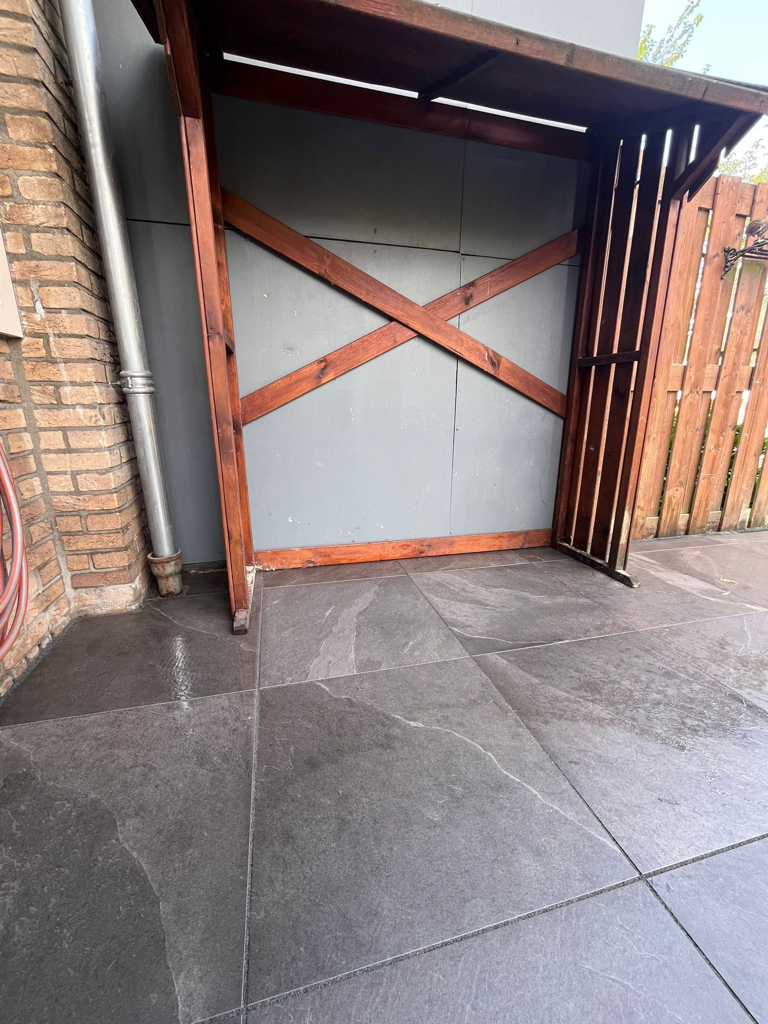 Houten afdak met grijze achtergrond en bruine dwarsbalken, op een terras met zwarte tegels.