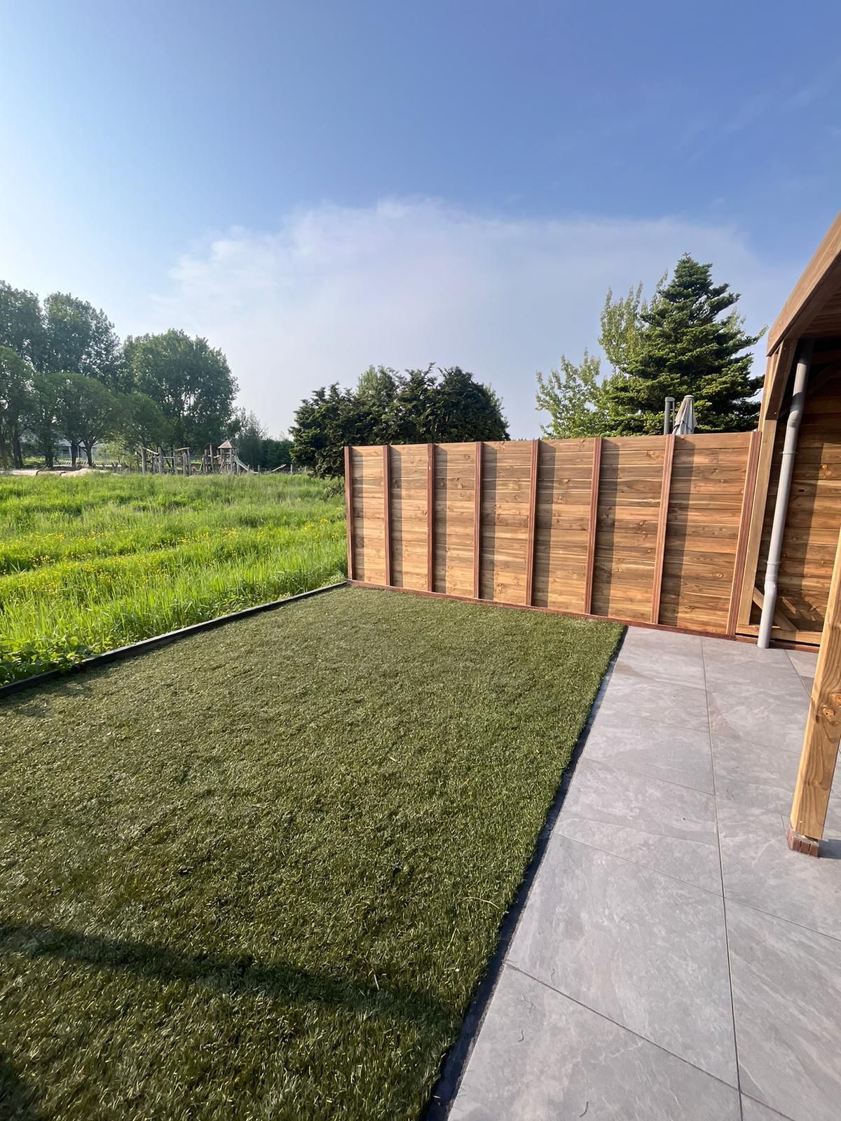 Houten schutting naast kunstgras, met bomen op de achtergrond onder een blauwe hemel.
