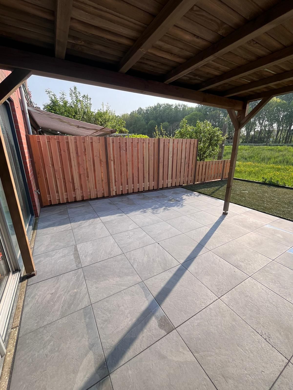 Terras met grijze natuurstenen tegels onder een houten pergola, dat uitkomt op een houten schutting en een groen veld.